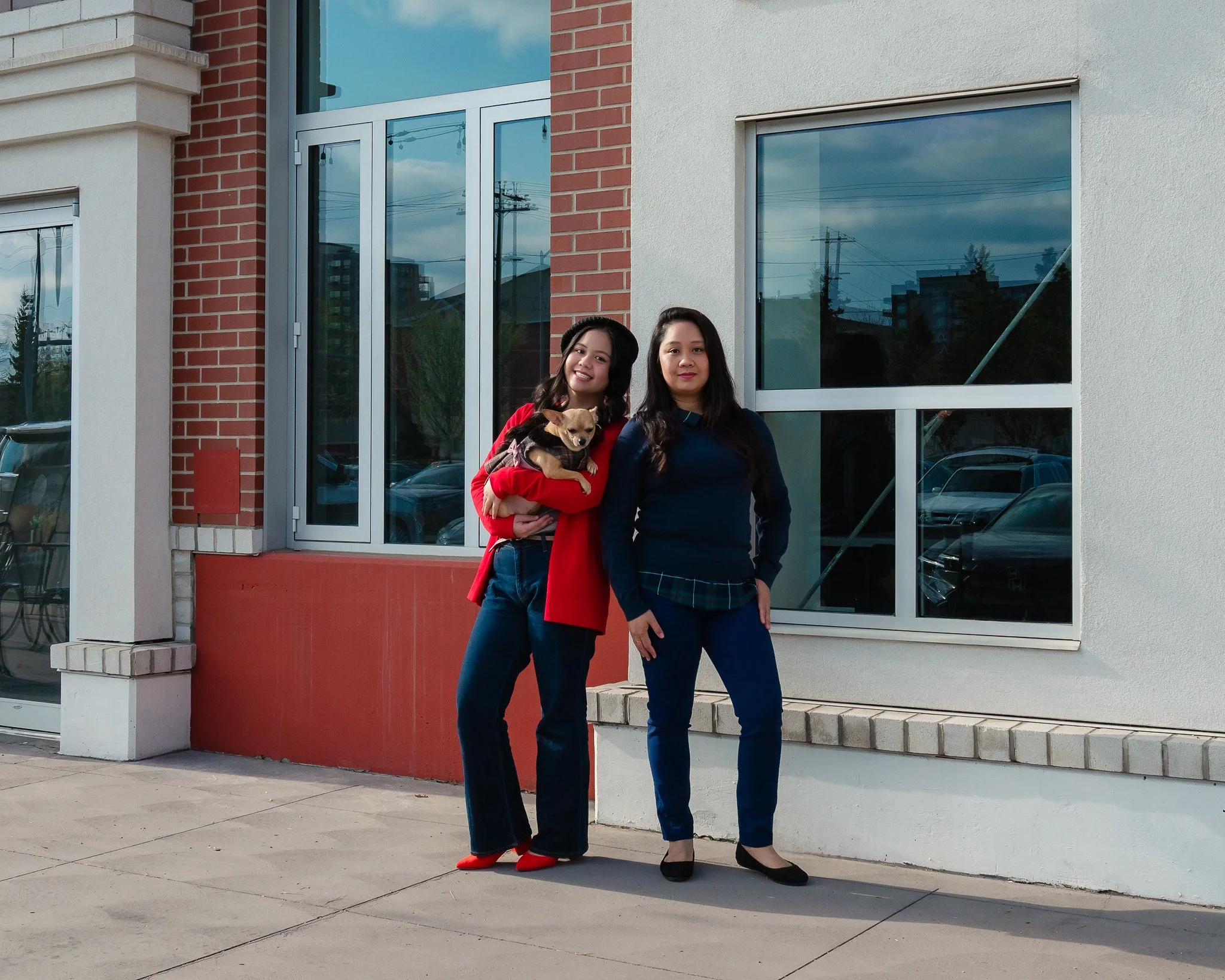 Two women standing in front of a modern building with large windows. One woman is holding a small dog. The woman on the left has dark hair, wearing a red blazer and matching shoes, and is smiling. The woman on the right has dark hair, wearing a dark 