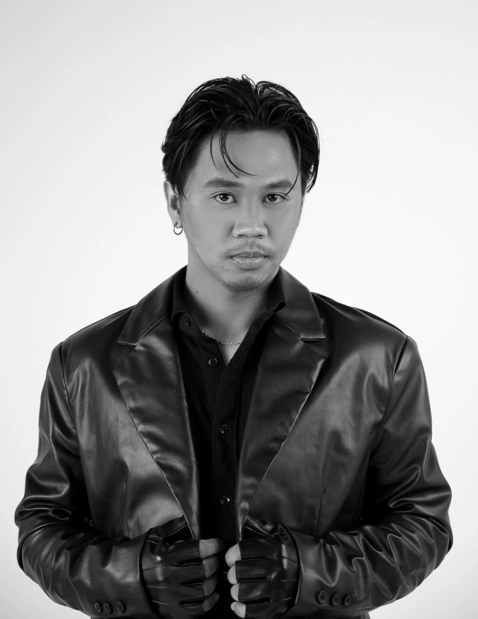 Black and white portrait of a man with dark hair styled back, wearing hoop earrings, a leather jacket, and gloves, with a serious expression against a plain background.