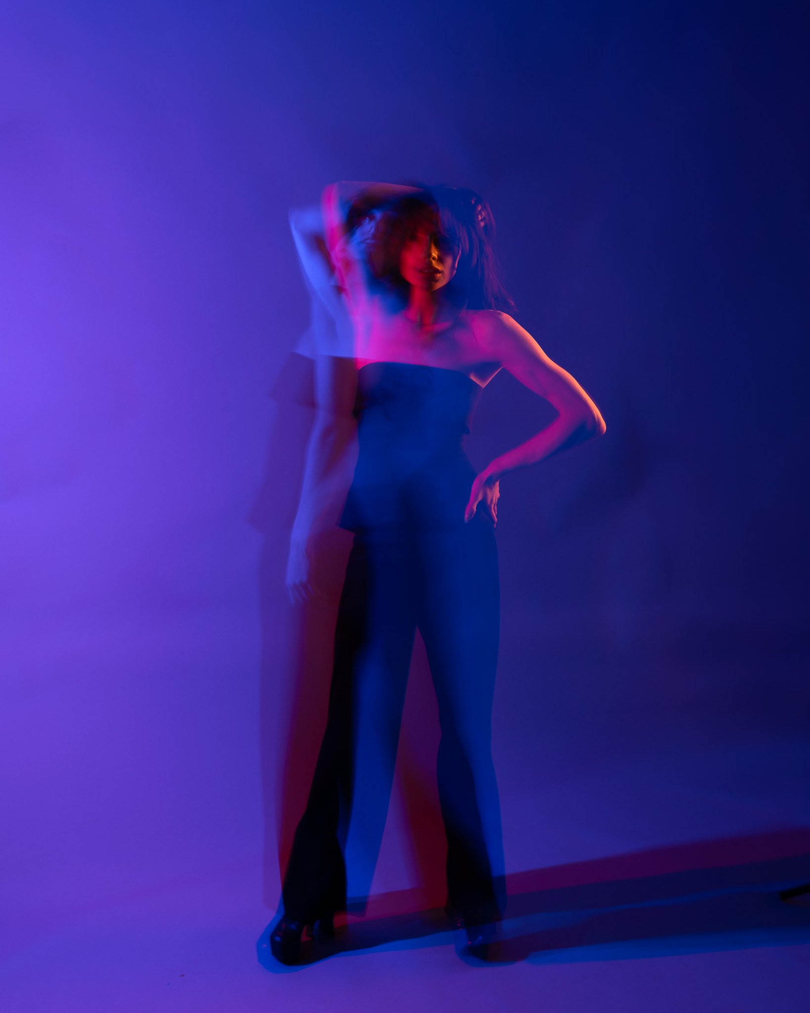 A woman with dark hair is posing in front of a dark background lit with vibrant purple and blue lights. She is wearing a strapless black top and black pants, with one hand on her hip and the other behind her head. The motion blur effect creates multi