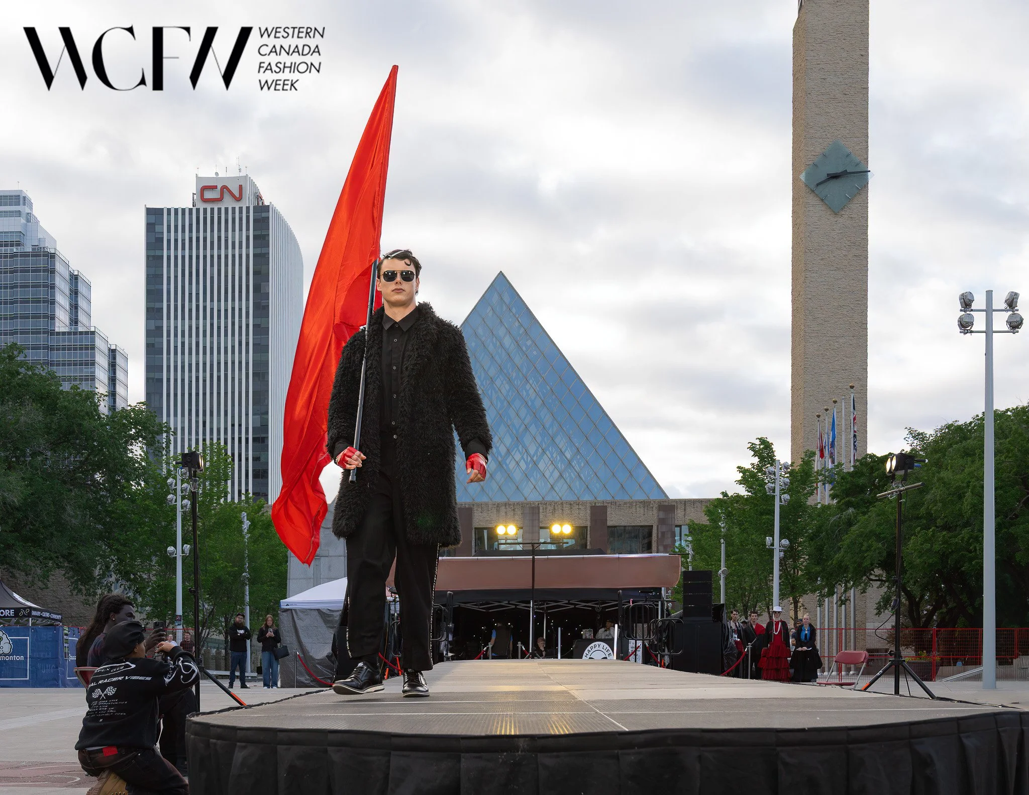 A performer dressed in black with sunglasses, red gloves, and a fur coat walks on an outdoor stage at Western Canada Fashion Week, with city skyscrapers and a cloudy sky in the background.