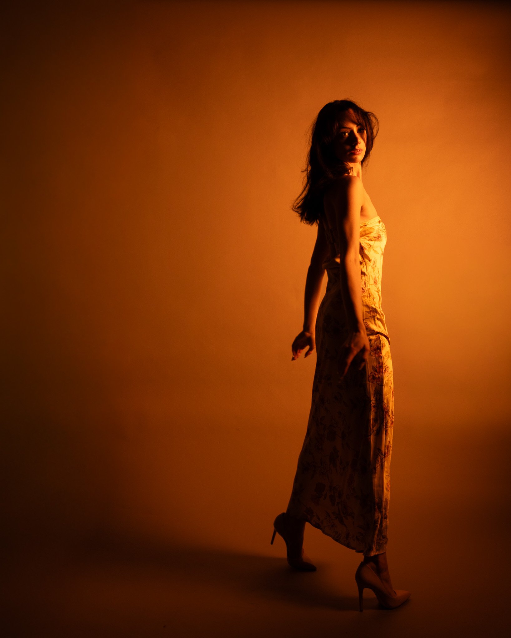 A woman standing on high heels in a silhouette against a warm orange background, wearing a long, patterned dress.