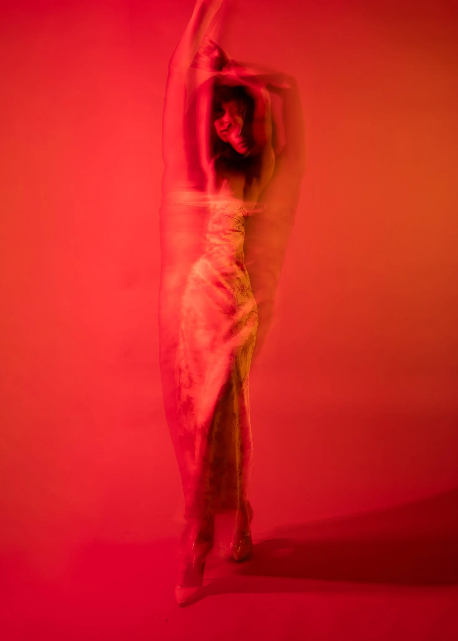 A woman with dark hair and a light-colored top and floral patterned pants stands against a red background, striking a dance pose with one arm raised above her head and the other bent behind her head.