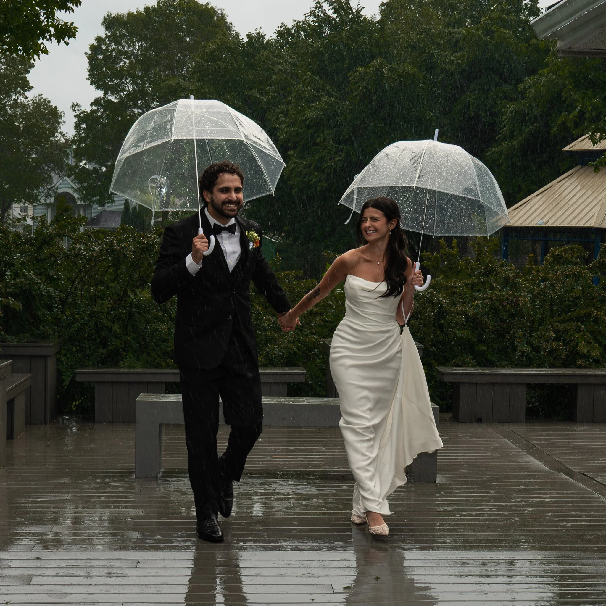 A smiling man in a tuxedo and a woman in a wedding dress walk hand in hand in the rain, each holding a transparent umbrella, on a wet outdoor wooden deck surrounded by greenery.