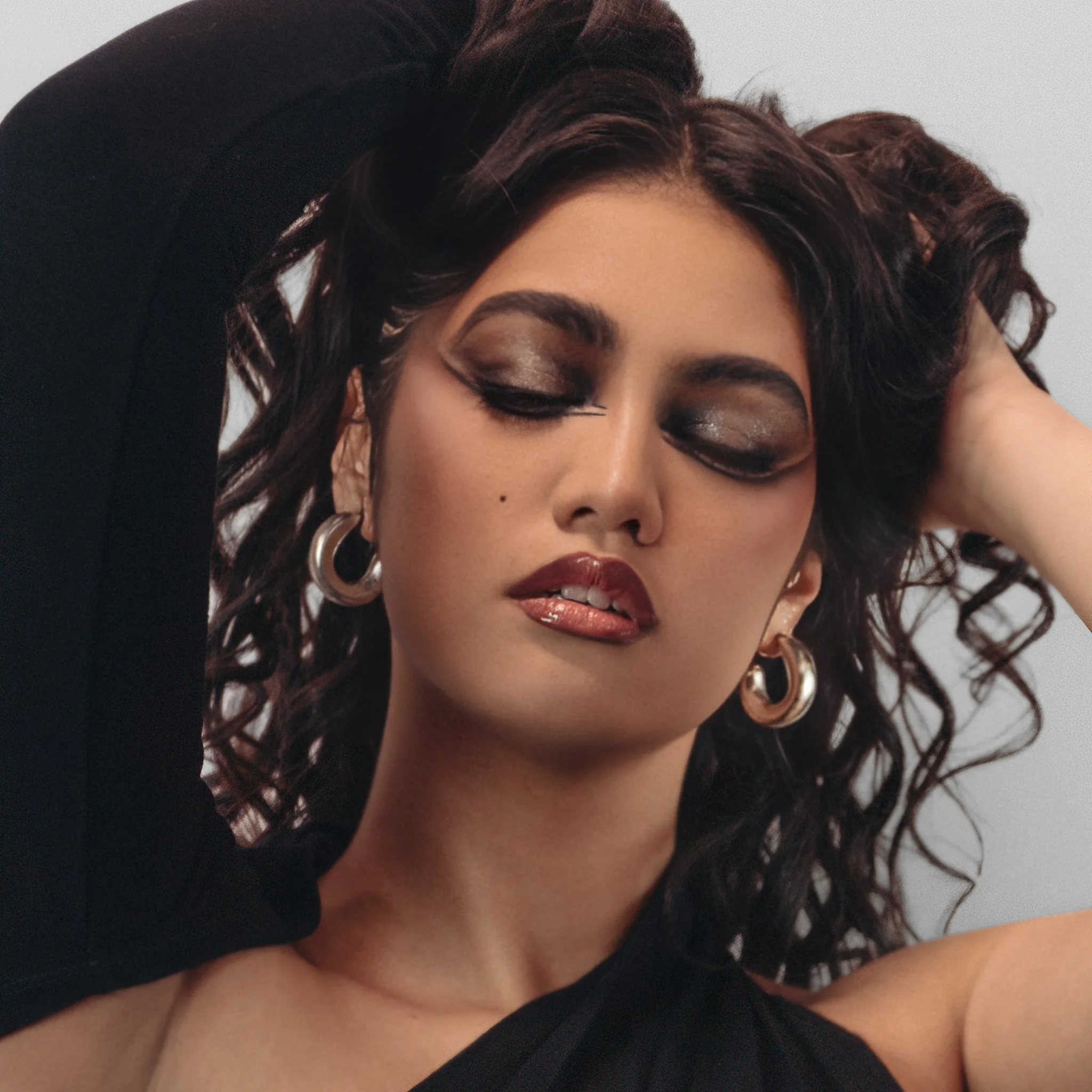 Close-up of a woman's face with makeup and large hoop earrings, with her eyes closed and one hand resting on her head.