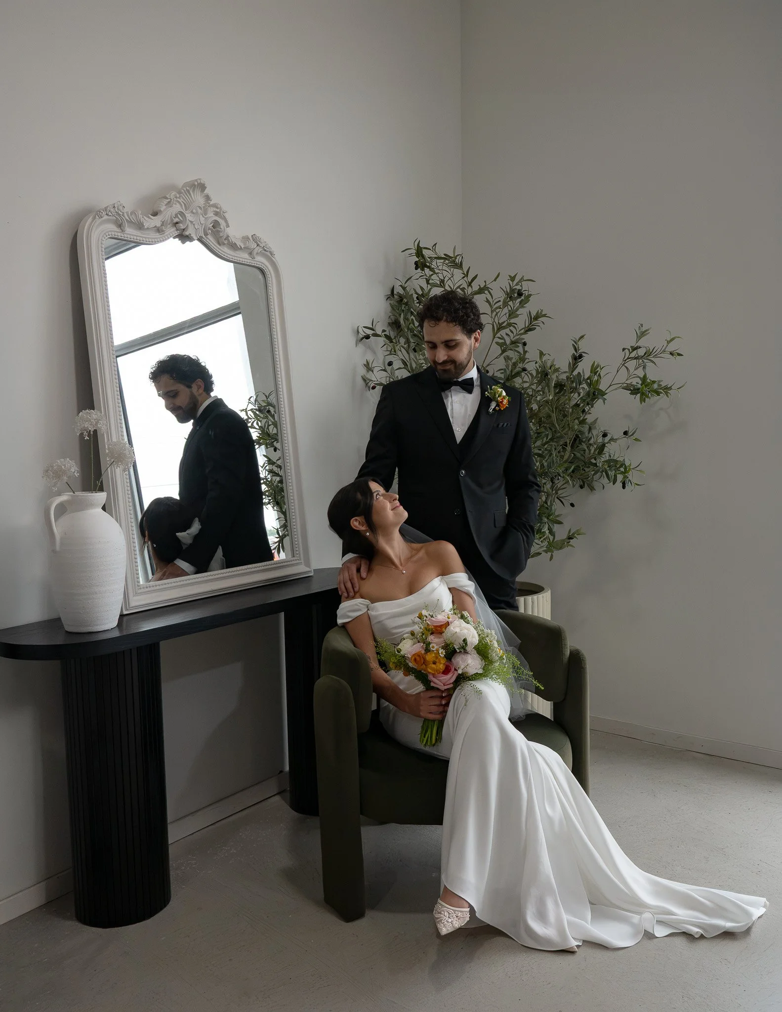 A bride sitting in a green chair holding a bouquet of flowers, while a groom in a tuxedo stands beside her, looking at her lovingly. They are in a room with a large mirror, a white vase, and some greenery.