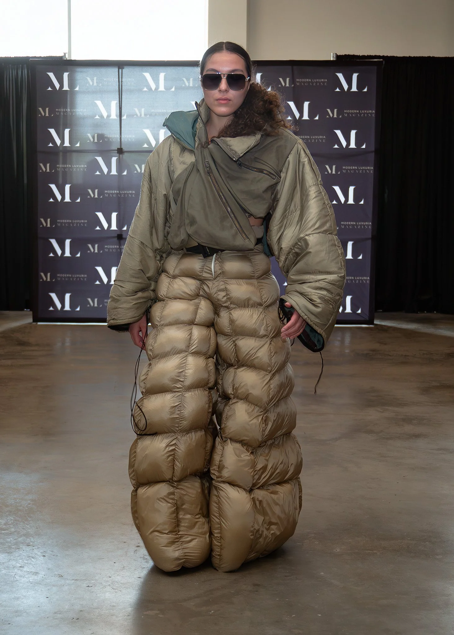 A woman wearing oversized sunglasses and a beige puffy jacket that extends to the floor, standing in front of a backdrop with white logo patterns on a black background at an indoor event.