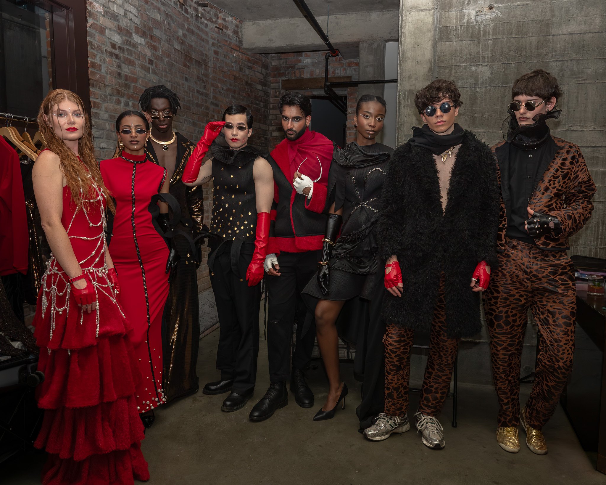 A group of eight diverse individuals dressed in fashionable and elaborate outfits poses together in an industrial-style room with brick and concrete walls.