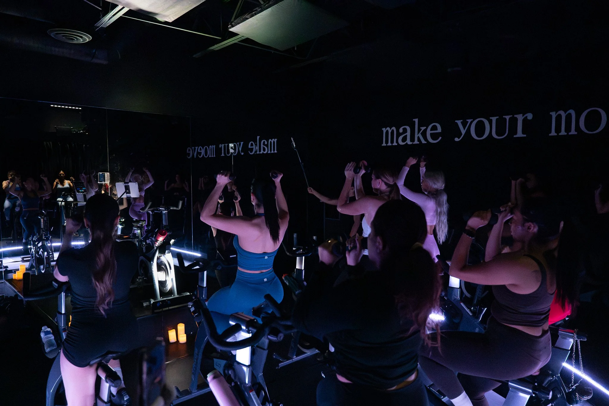 People working out on stationary bikes in a dark room with purple lighting, mirrors on the wall, and motivational text on the wall that reads 'make your move'.