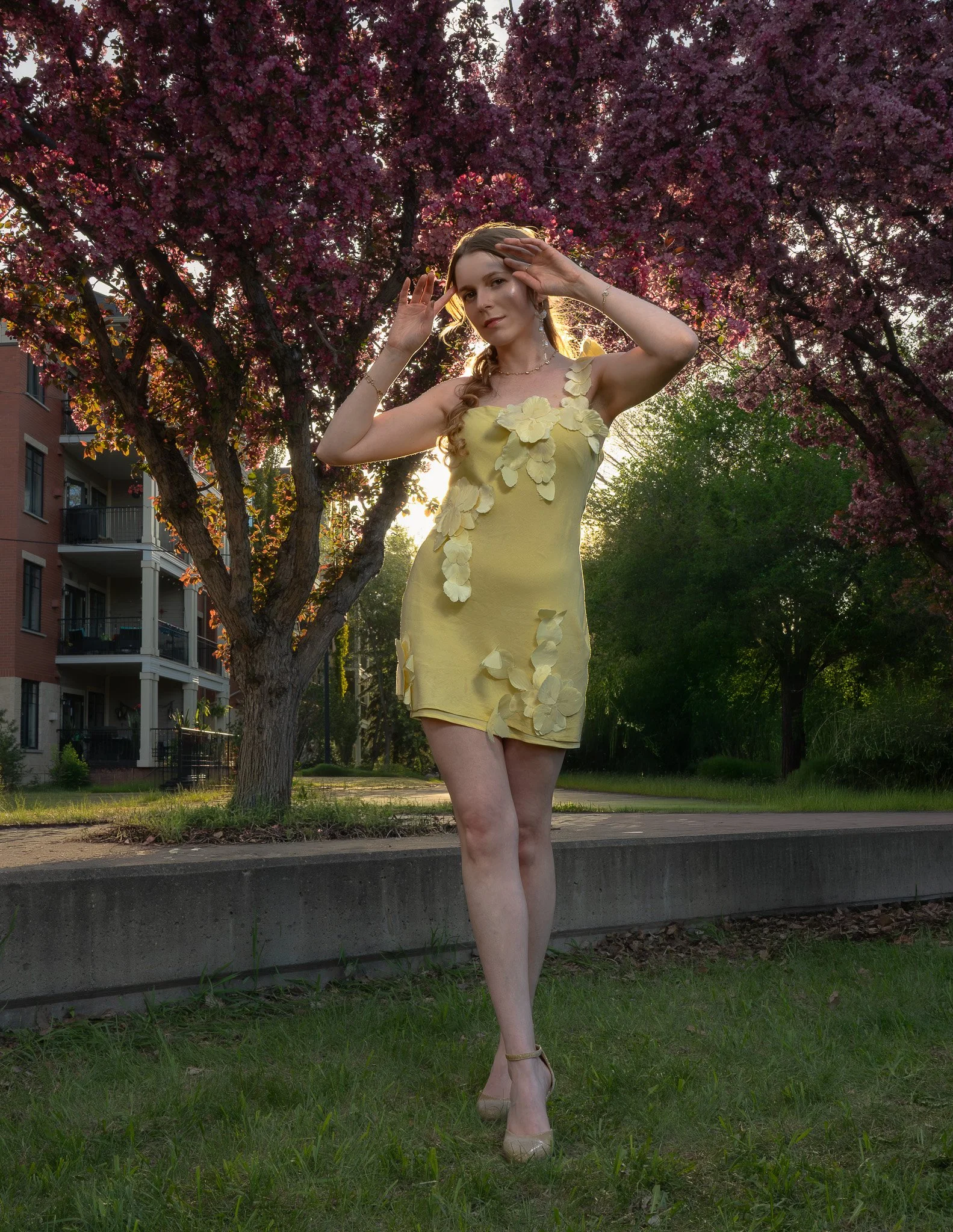 A woman in a yellow dress decorated with flower patterns, standing outdoors on a grassy area near a concrete edge, with trees and pink blossoms in the background, and apartment buildings on the left side of the image.