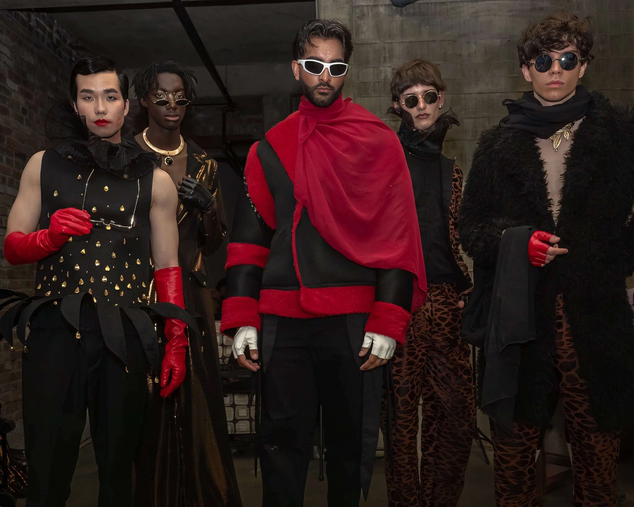 Group of five people dressed in fashionable, bold clothing and accessories, standing in front of industrial-style background.