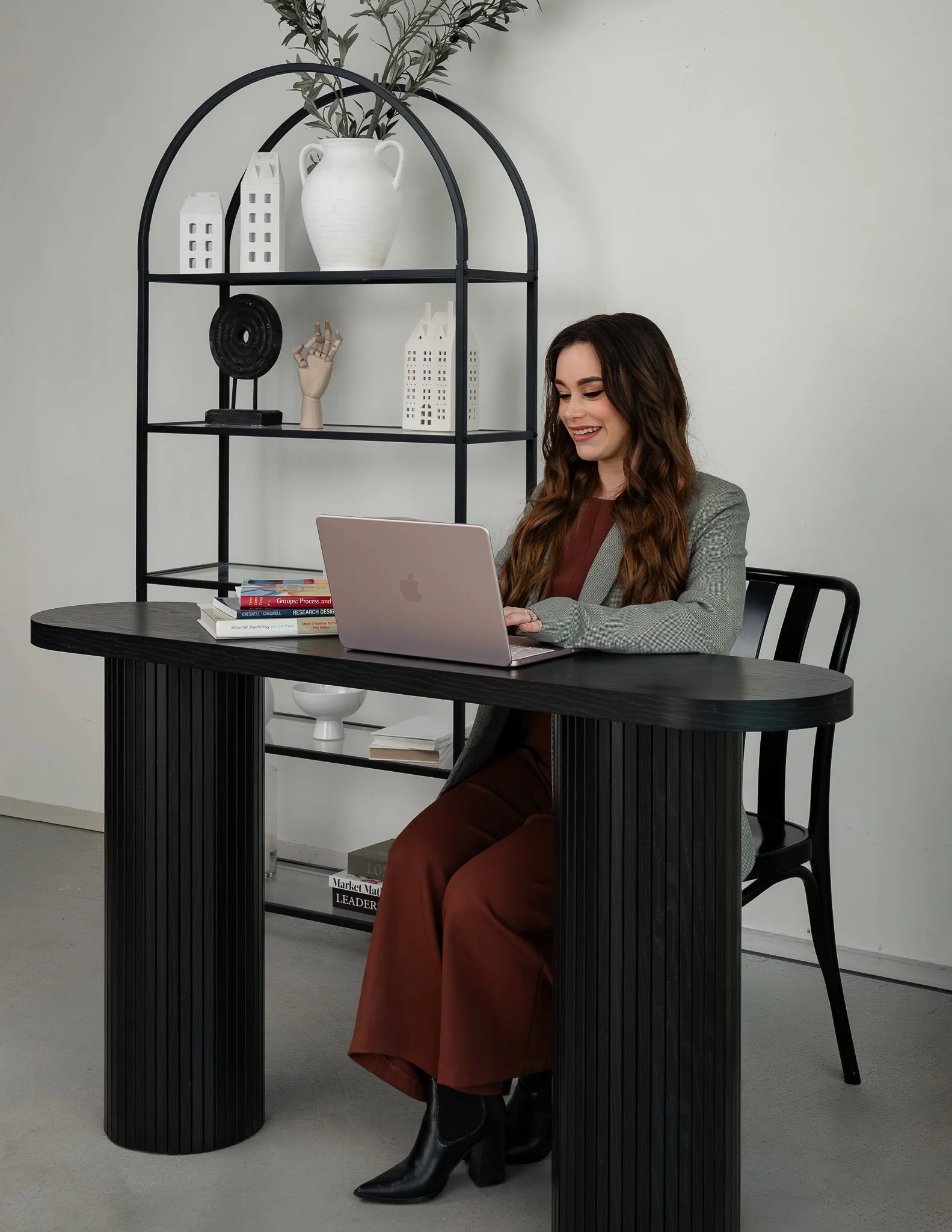 A young woman with long wavy hair sitting at a black table, smiling and looking at her laptop. She is wearing a gray blazer, rust-colored pants, and black boots. Behind her is a black metal shelving unit with decorative items, including white ceramic