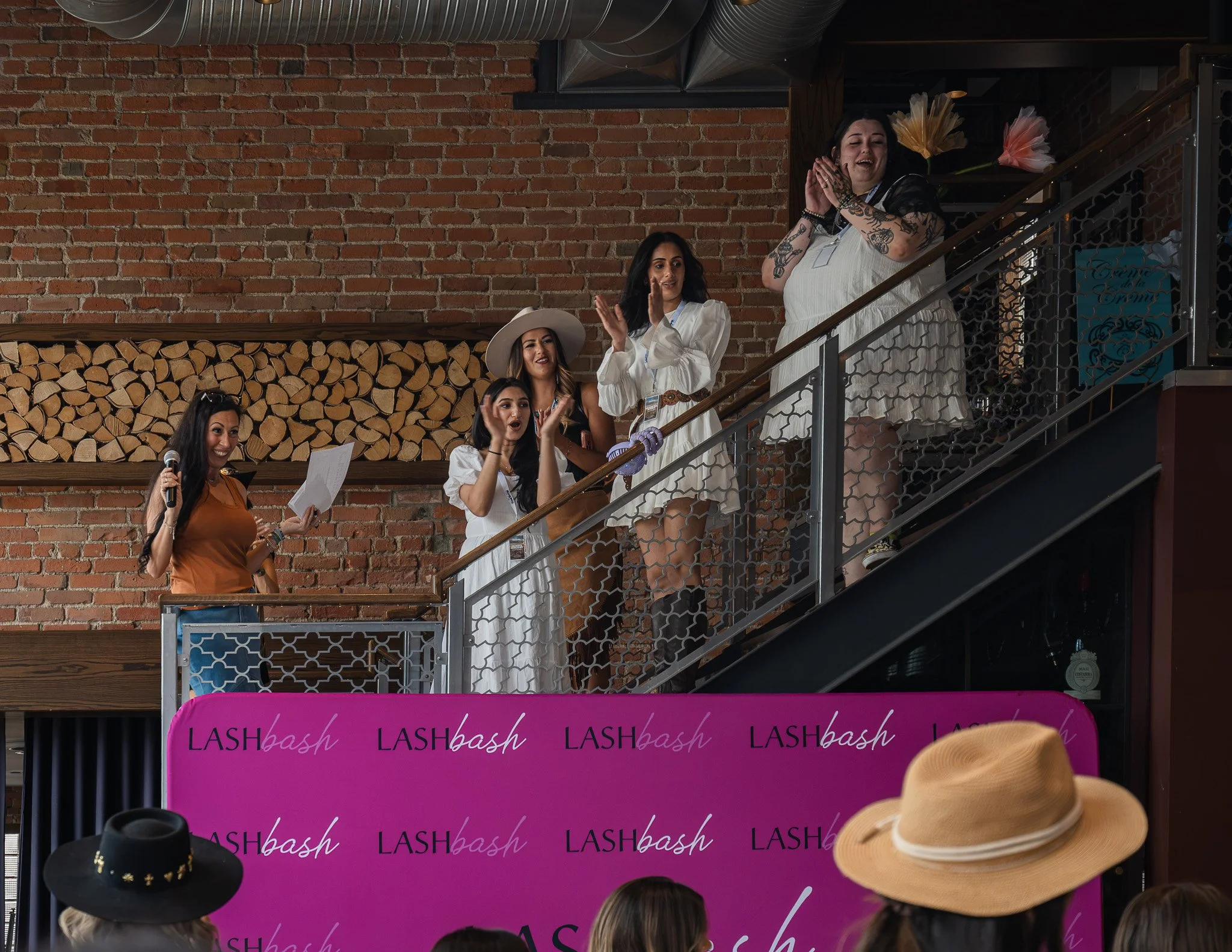 Group of women on a stage observing a beauty or fashion event, with a large pink banner in the foreground and a brick wall background.
