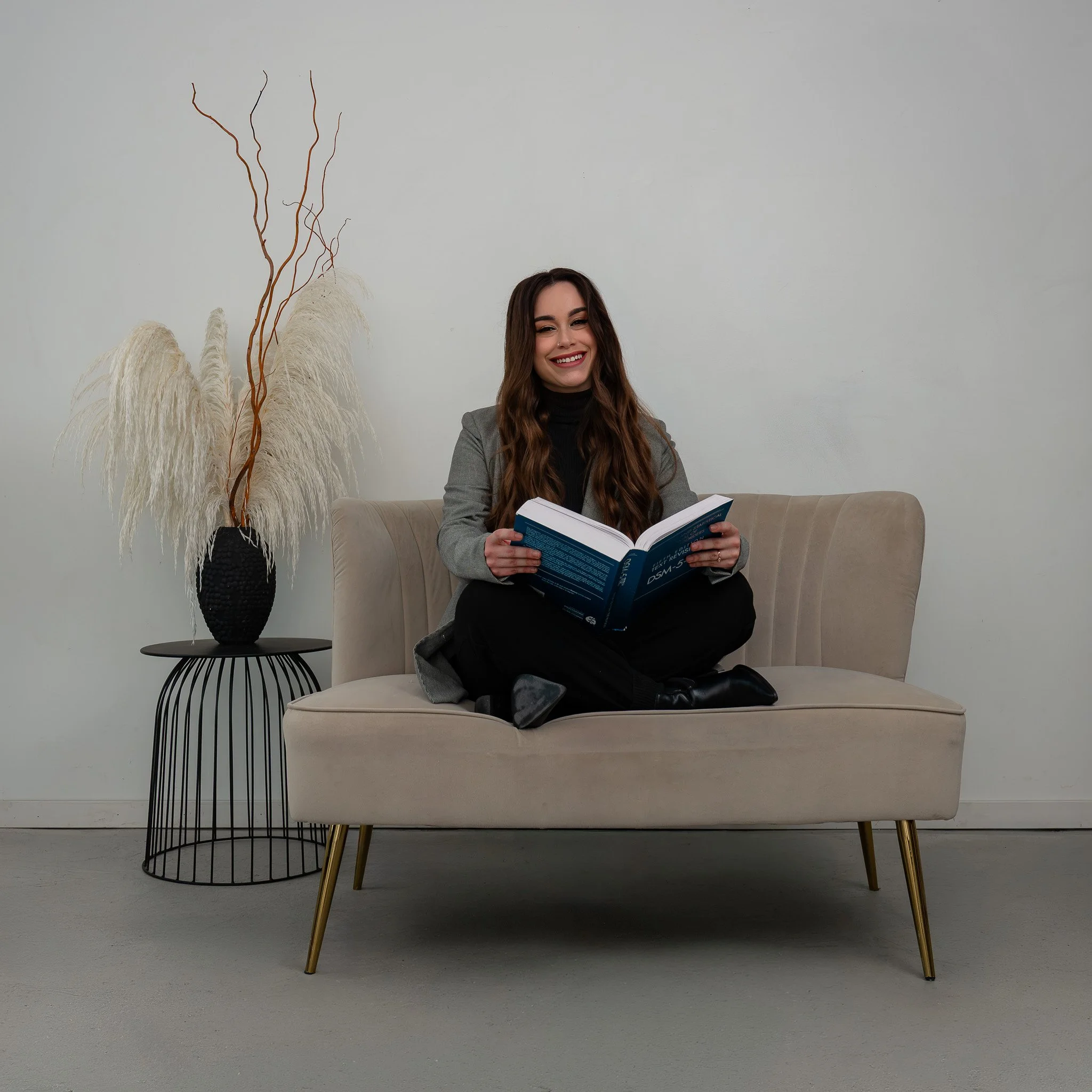 A woman with long brown hair, smiling, sitting on a beige sofa, reading a book, dressed in a gray blazer and black top, with a black shoe visible in her lap. Behind her, there's a white wall with decorative brown twigs and white pampas grass in a bla