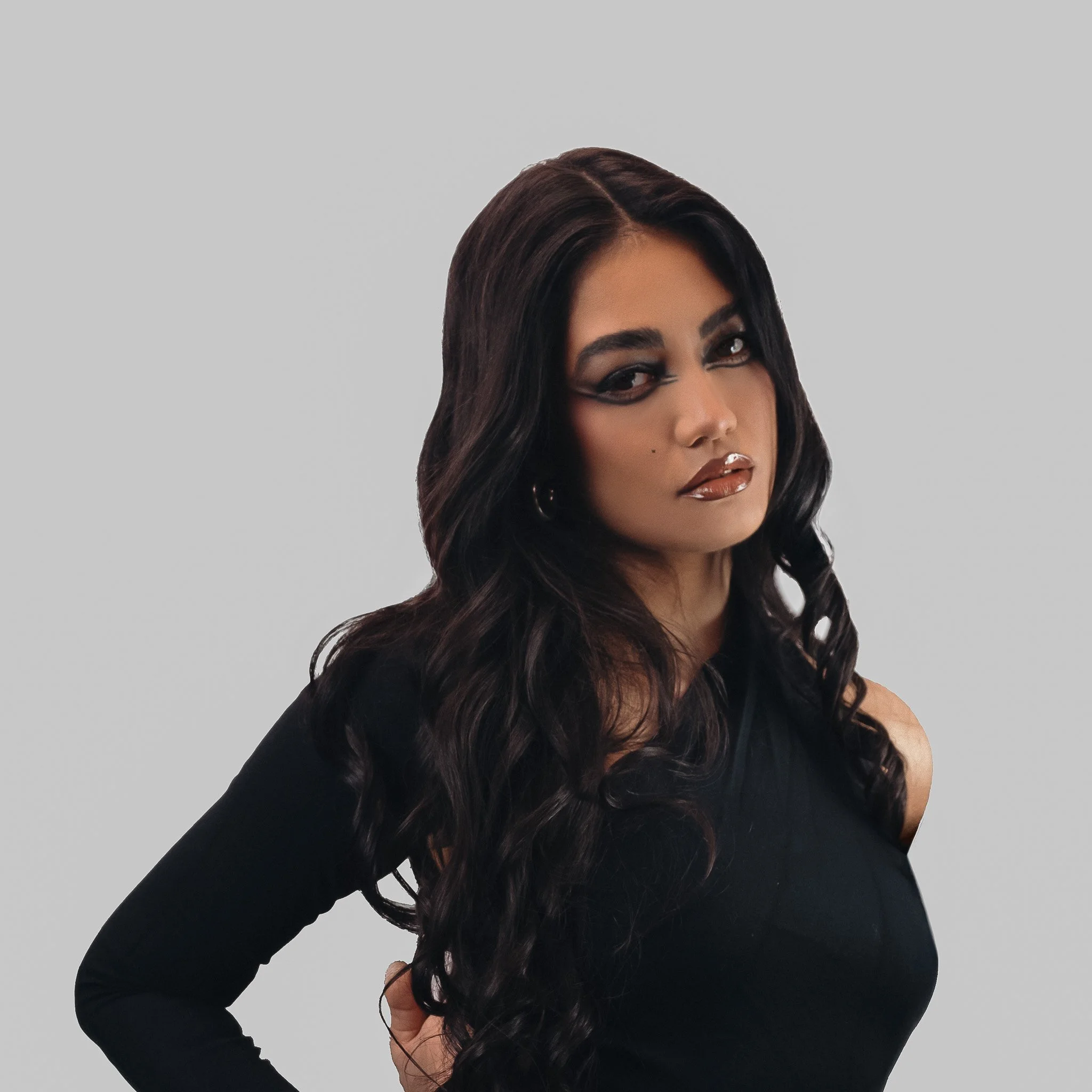 Close-up of a woman with long, wavy dark hair, wearing makeup with dark eye makeup and glossy lipstick, dressed in black, posing against a plain gray background.