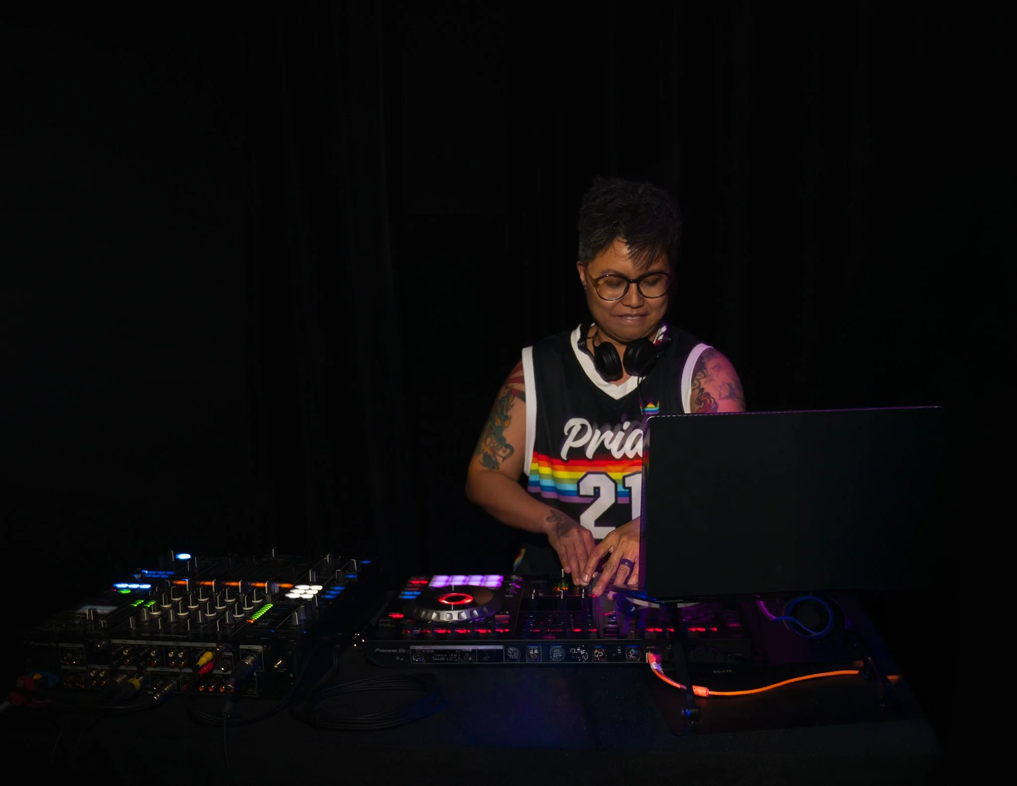 A woman with tattoos, short hair, glasses, and wearing a pride rainbow tank top is DJing with a Pioneer DJ controller in a dark setting.