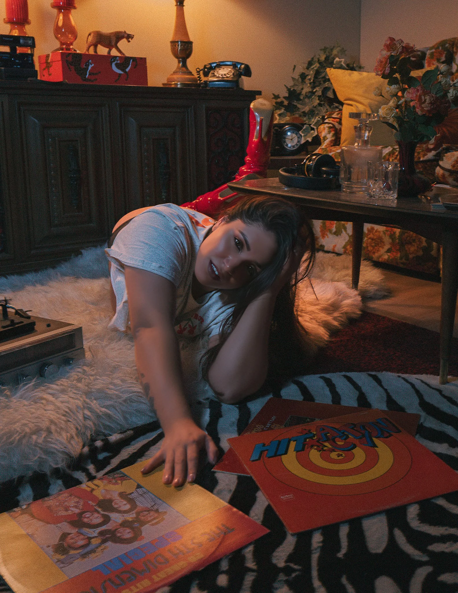 A young woman lying on the floor surrounded by vinyl record albums and a tabletop with a plant, a glass pitcher, and a figurine. The background contains a wooden cabinet with decorative items and a floral-patterned couch with pillows.