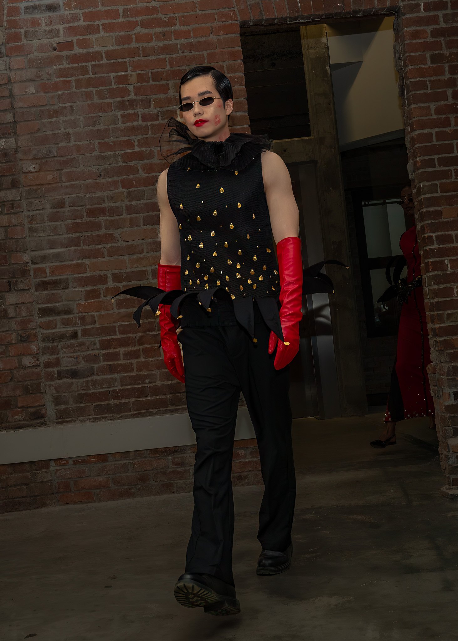Person dressed in black with a ruffled collar, red gloves, black pants, and sunglasses standing indoors against a brick wall, with another person in red clothing visible in the background.