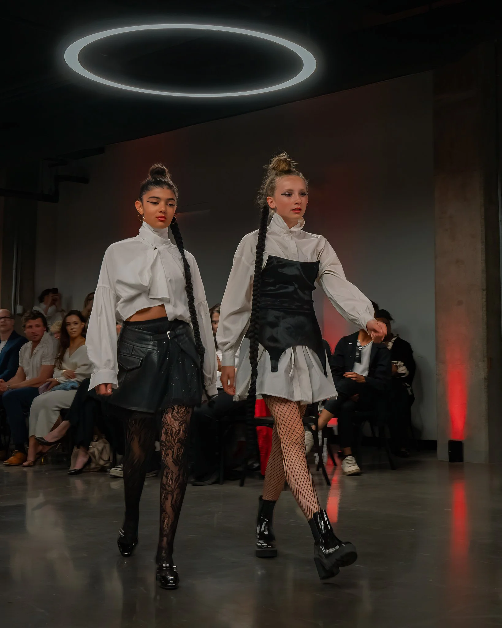 Two female models walk in a fashion show, wearing black and white outfits with fishnet stockings, platform shoes, and high ponytail hairstyles, with audience members watching in the background and a circular ceiling light above.