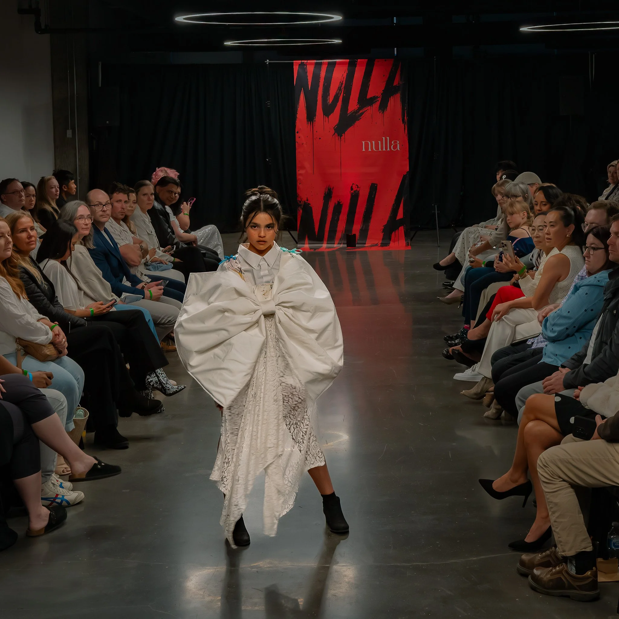 A fashion runway show with a model walking down the runway in a white lace dress and black boots, audience members seated on either side, a red and black banner in the background with the words 'NULLA' multiple times.