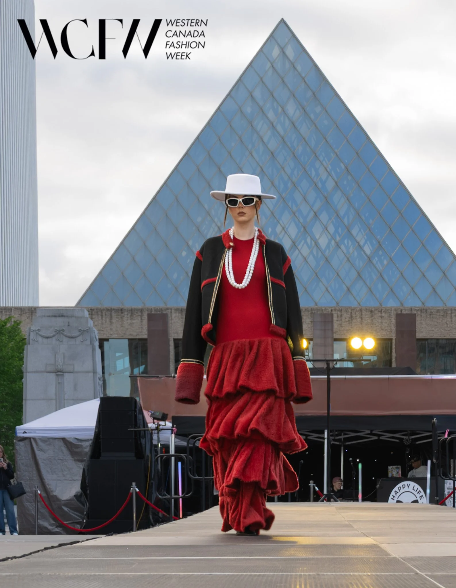 A woman walking on a runway during Western Canada Fashion Week, wearing a red layered textured skirt, matching red top, black coat with red accents, large white hat, white sunglasses, and pearl necklaces, with a modern glass building in the backgroun