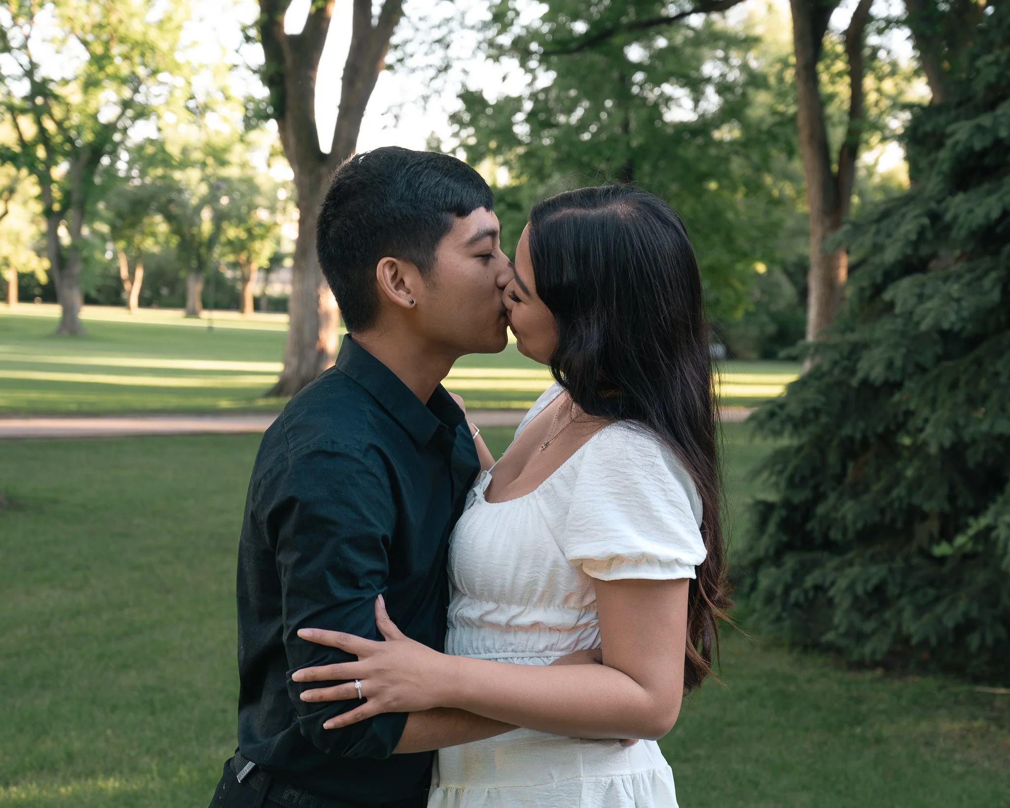 A couple kissing outdoors in a park with green trees in the background.
