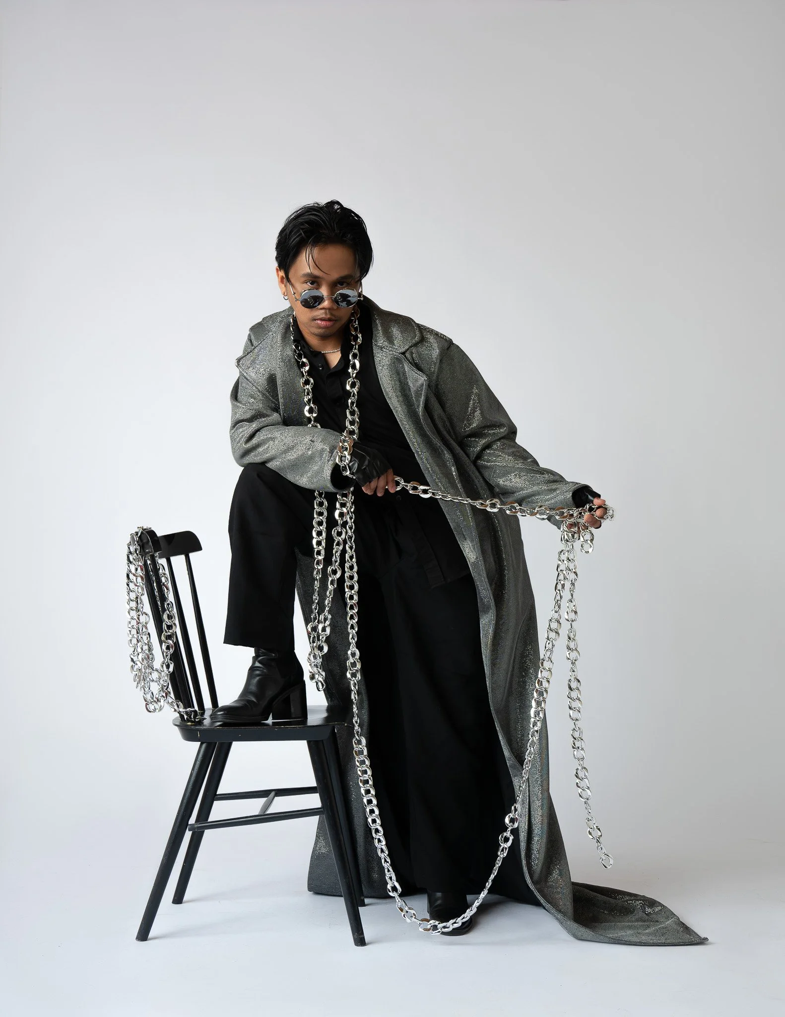 A man with black hair, wearing sunglasses, a metallic silver coat, black outfit, and multiple chunky silver chains, is sitting on a black chair and holding a silver chain. The background is plain white.