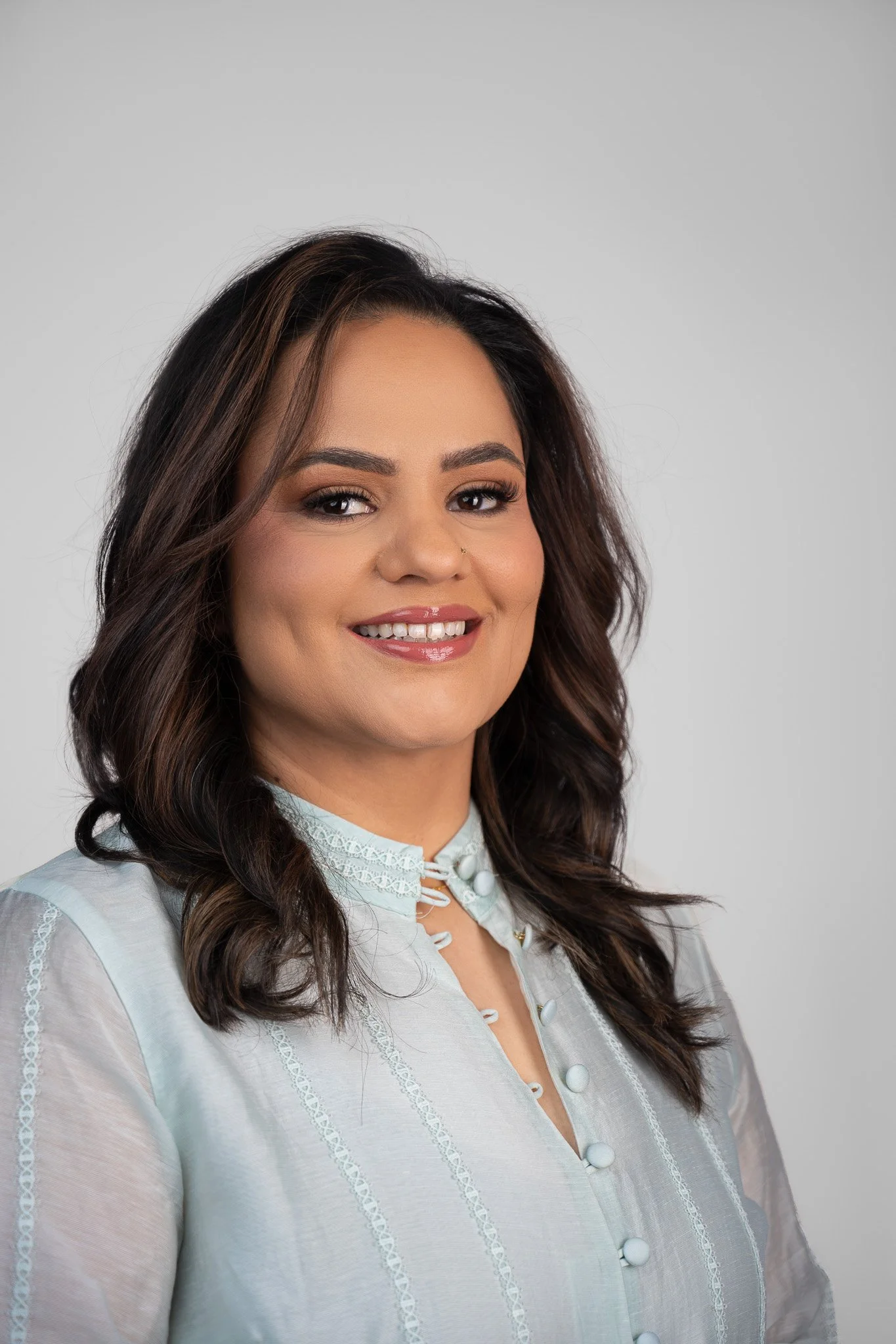 A woman with wavy dark brown hair and light makeup, wearing a sheer white blouse with embroidered details and pearl buttons, smiling against a plain light gray background.