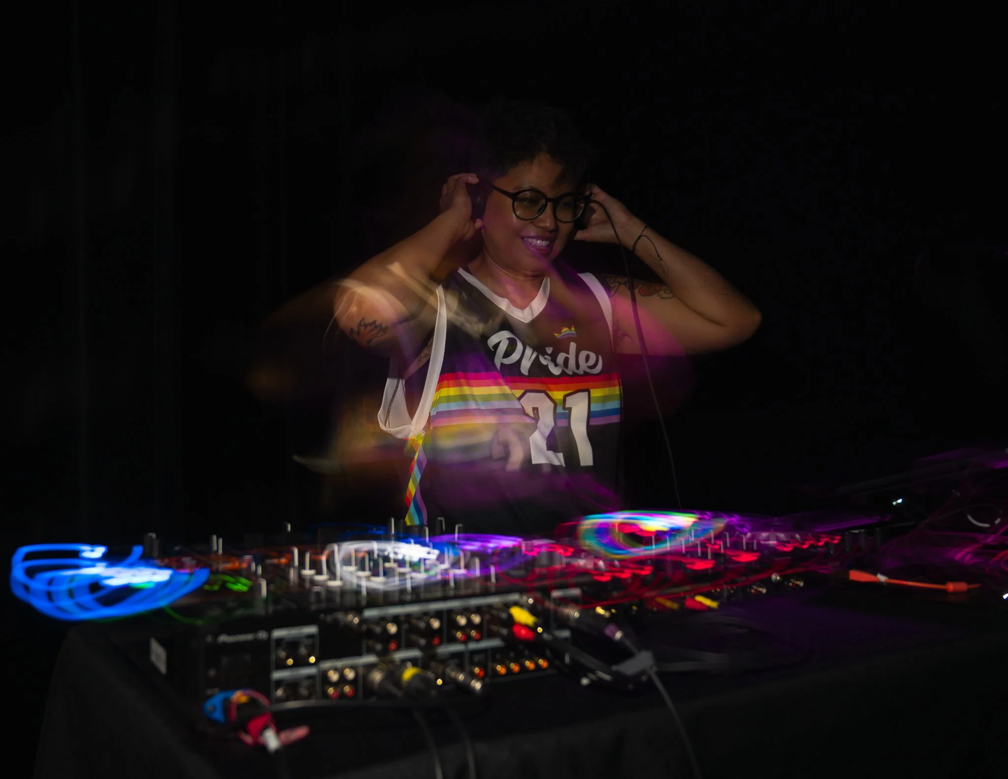 A person with short curly hair, glasses, and tattoos on arms, wearing a rainbow pride shirt, smiling while DJing at a music event with colorful lights and DJ equipment.
