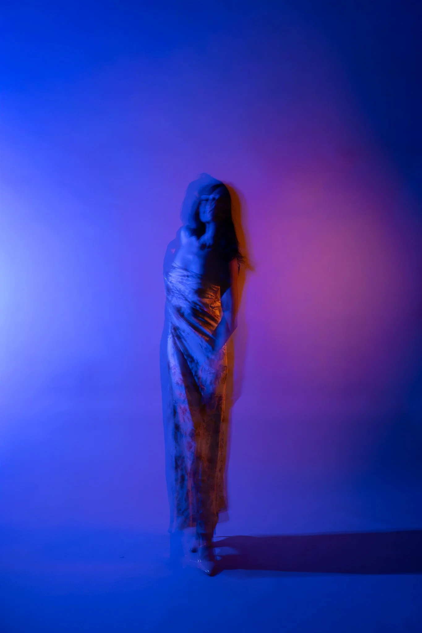 A woman with long hair standing against a wall illuminated with colorful blue, purple, and orange lights, wearing a sleeveless top and loose pants.