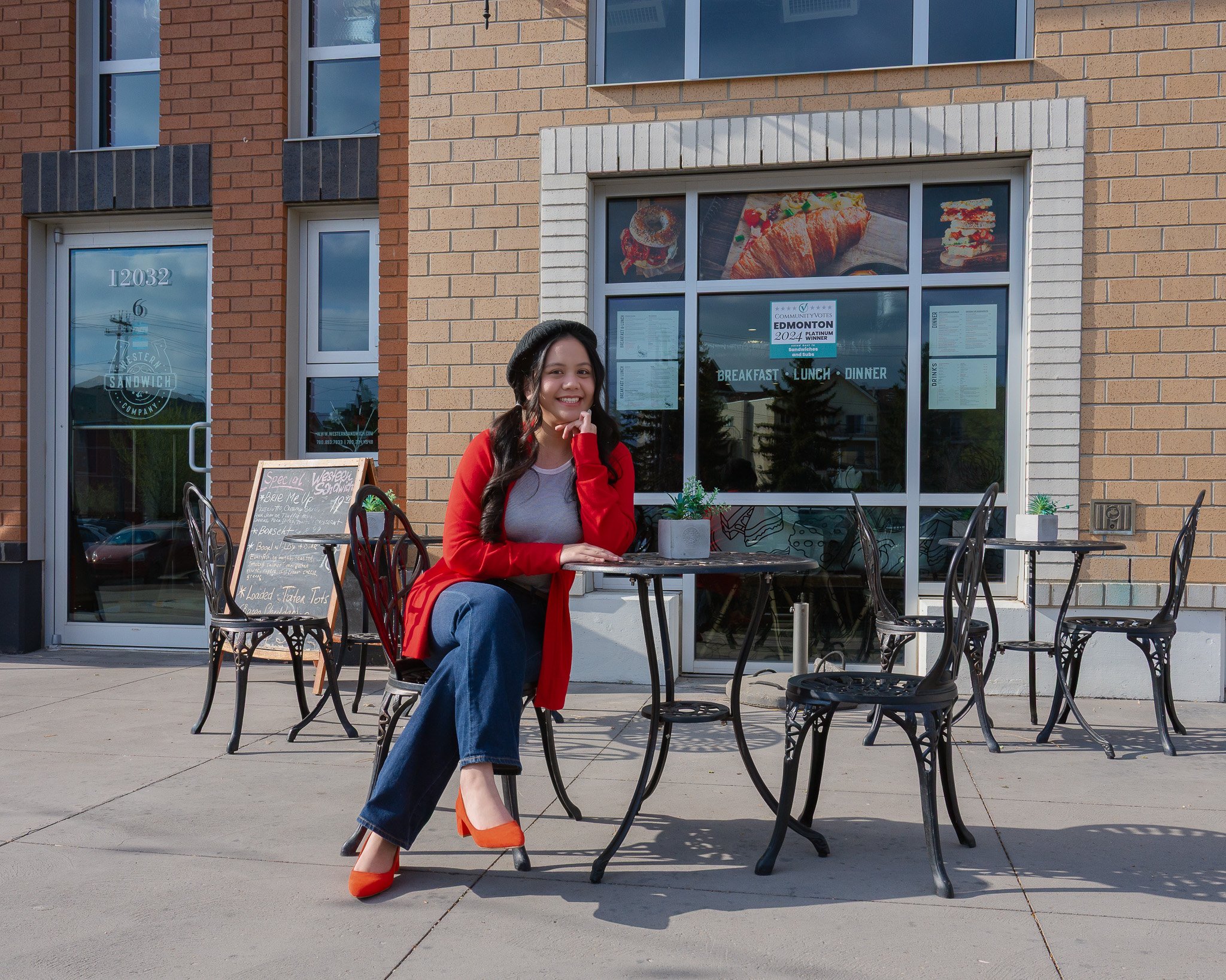 A woman sitting outside a restaurant with large windows, smiling and wearing a red jacket, gray top, blue jeans, and orange high heels, with a menu board and outdoor seating visible in the scene.