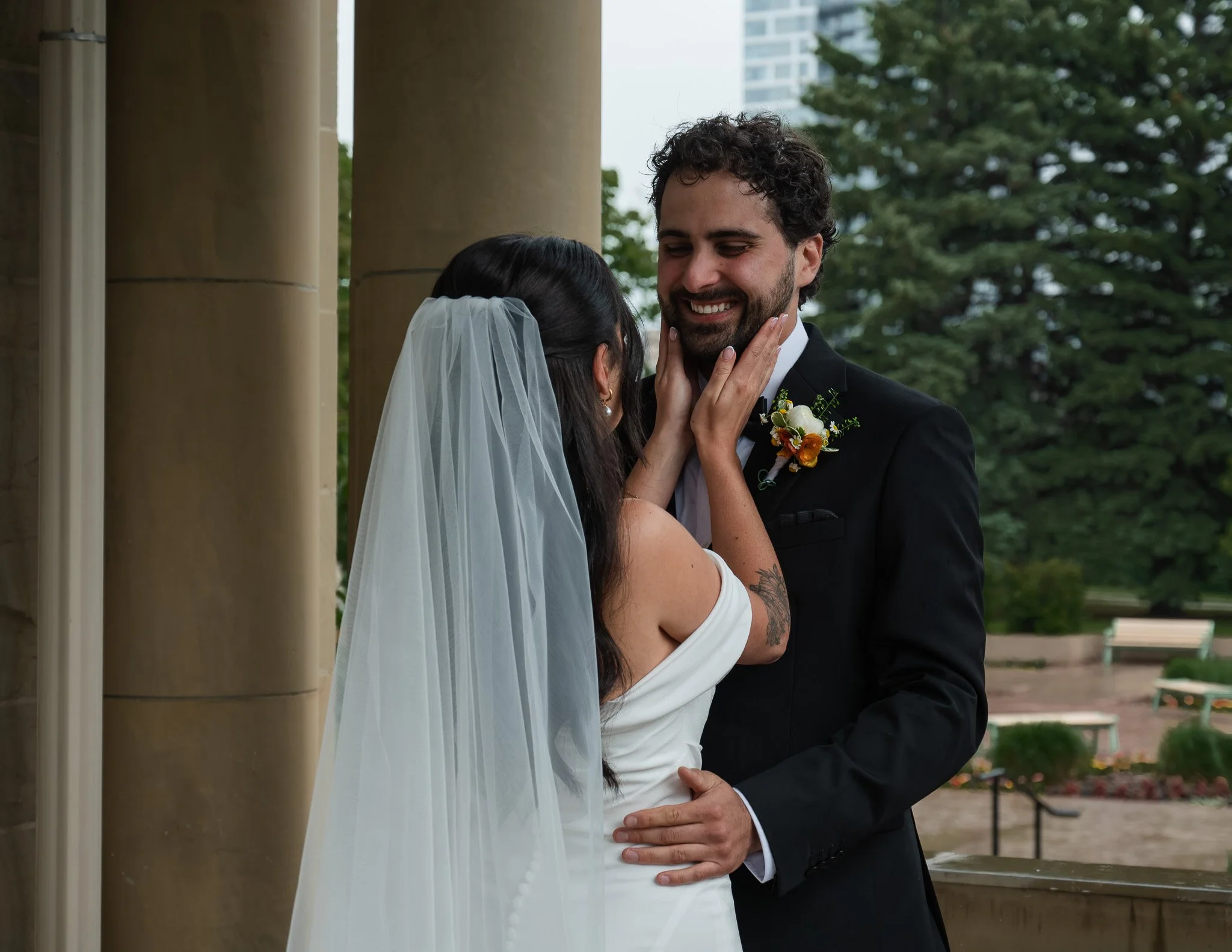 A bride and groom share a loving moment outdoors during their wedding ceremony, with trees and a building in the background.