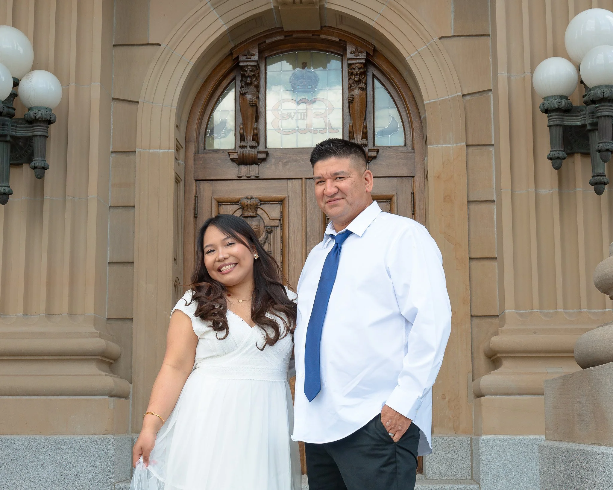 A man and woman standing in front of a large wooden door with decorative carvings and stained glass. The woman is smiling and wearing a white dress, and the man is wearing a white shirt with a blue tie.