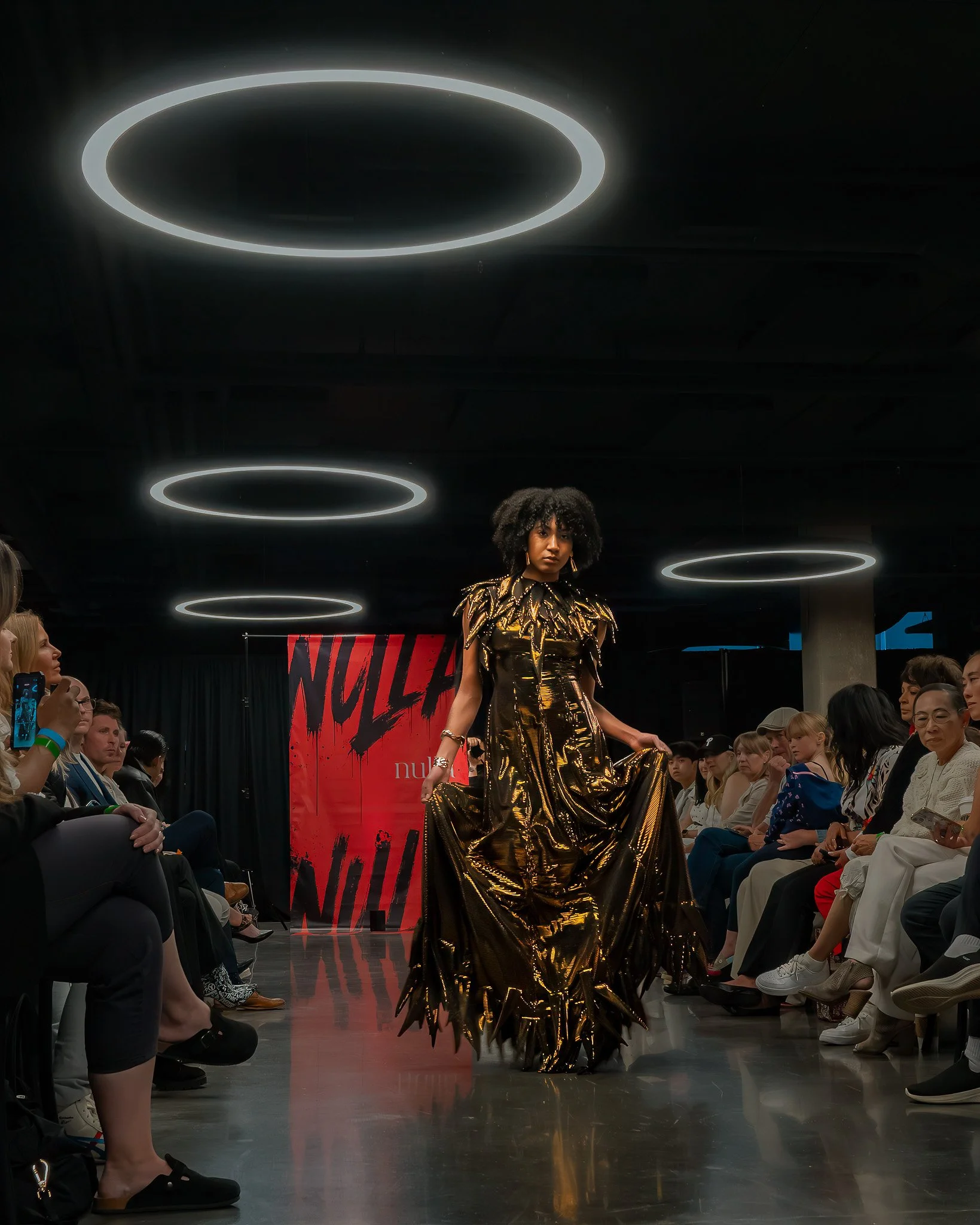 Fashion runway show with model in metallic gold dress and audience seated on both sides. Overhead modern circular lighting fixtures and a red banner with black and red writing.