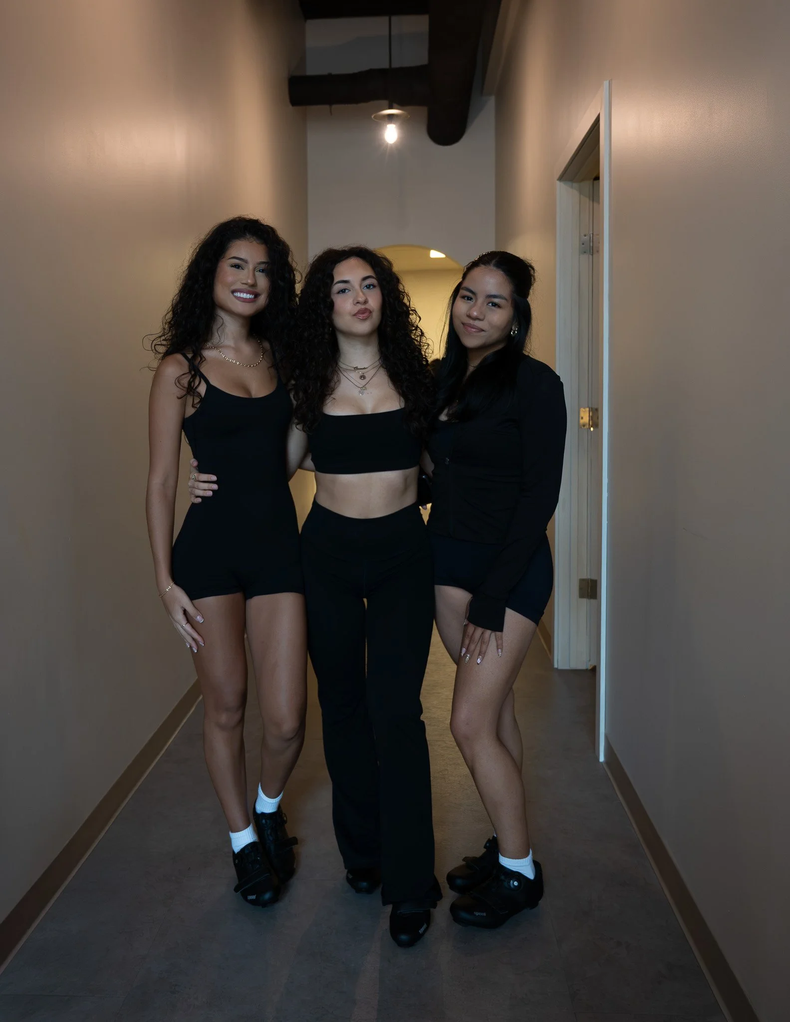 Three women standing in an indoor hallway, wearing black outfits and roller skates, smiling at the camera.