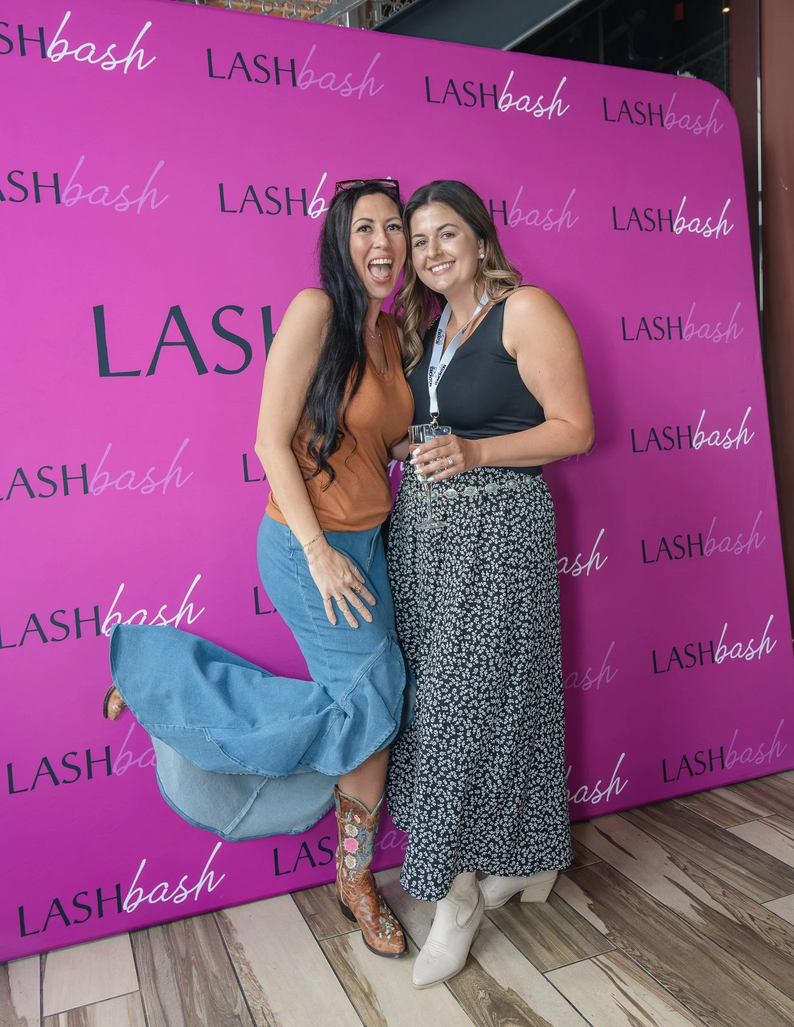 Two women standing together in front of a pink background with the words 'LASHbash' repeated across it. One woman is wearing a brown top, blue jeans, and cowboy boots with floral embroidery. The other woman is wearing a black top and a patterned skir