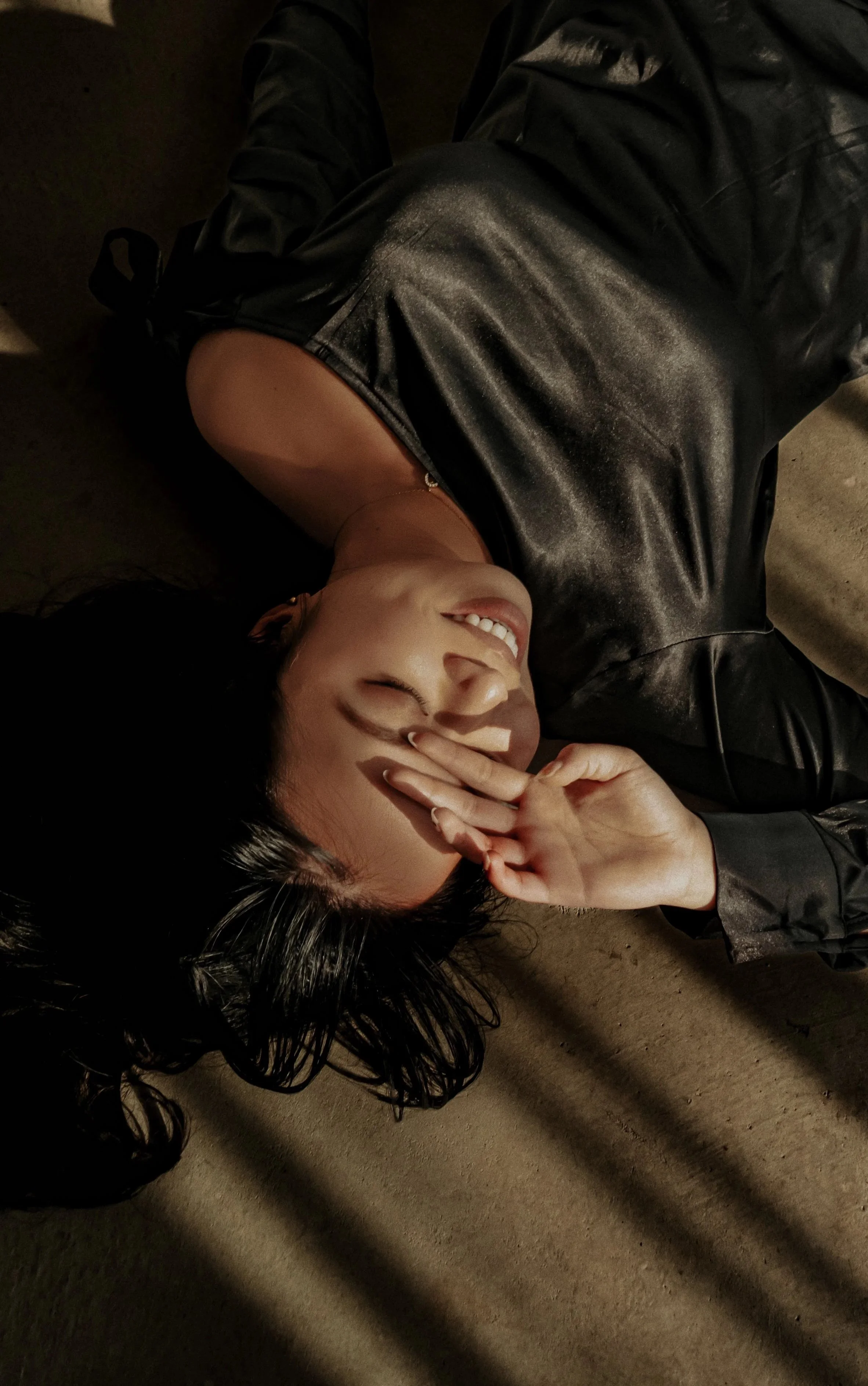A woman with dark hair lying on the ground, wearing a black leather jacket, smiling, with her hand near her face showing a peace sign.