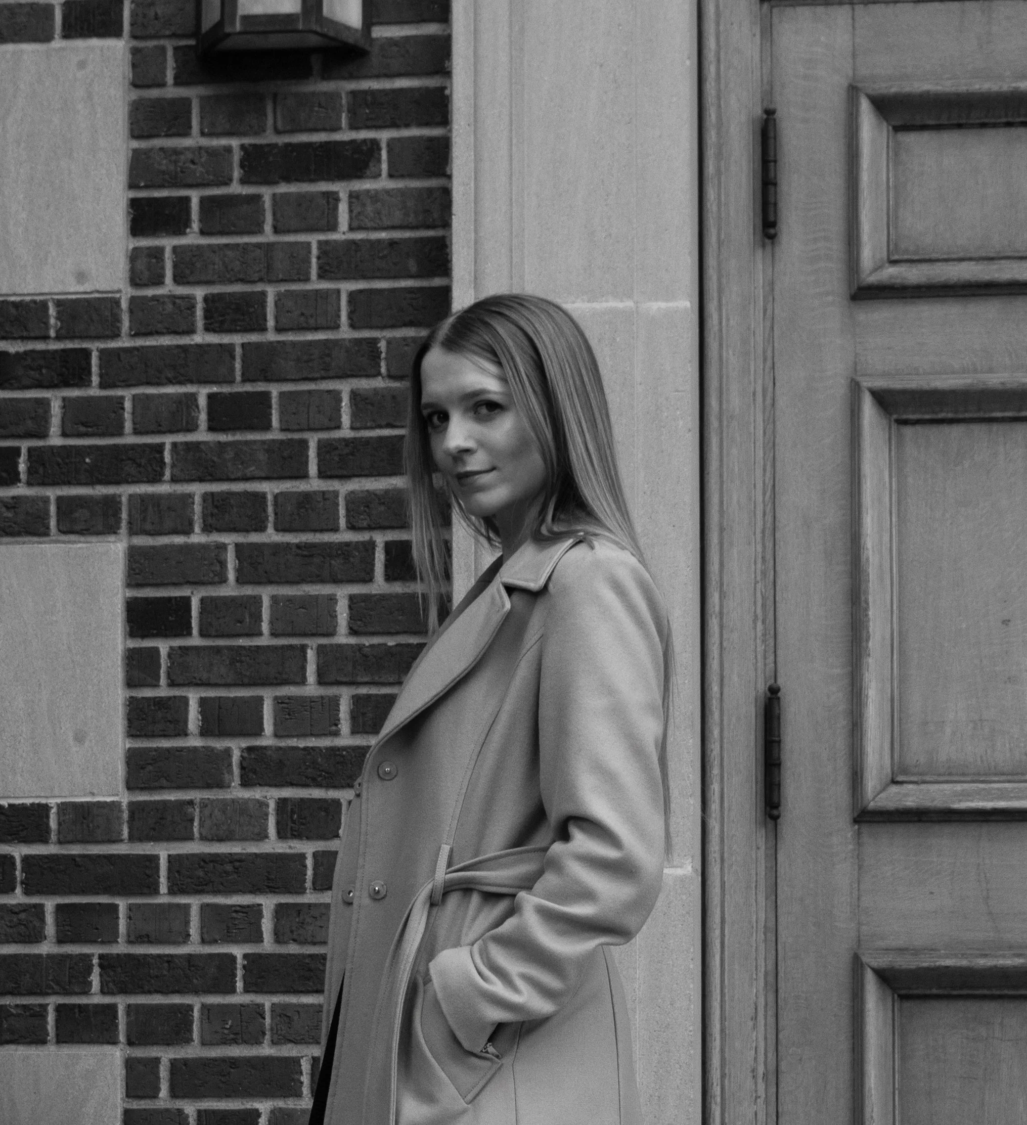 A young woman with straight hair wearing a trench coat, standing against a brick wall and wooden door.