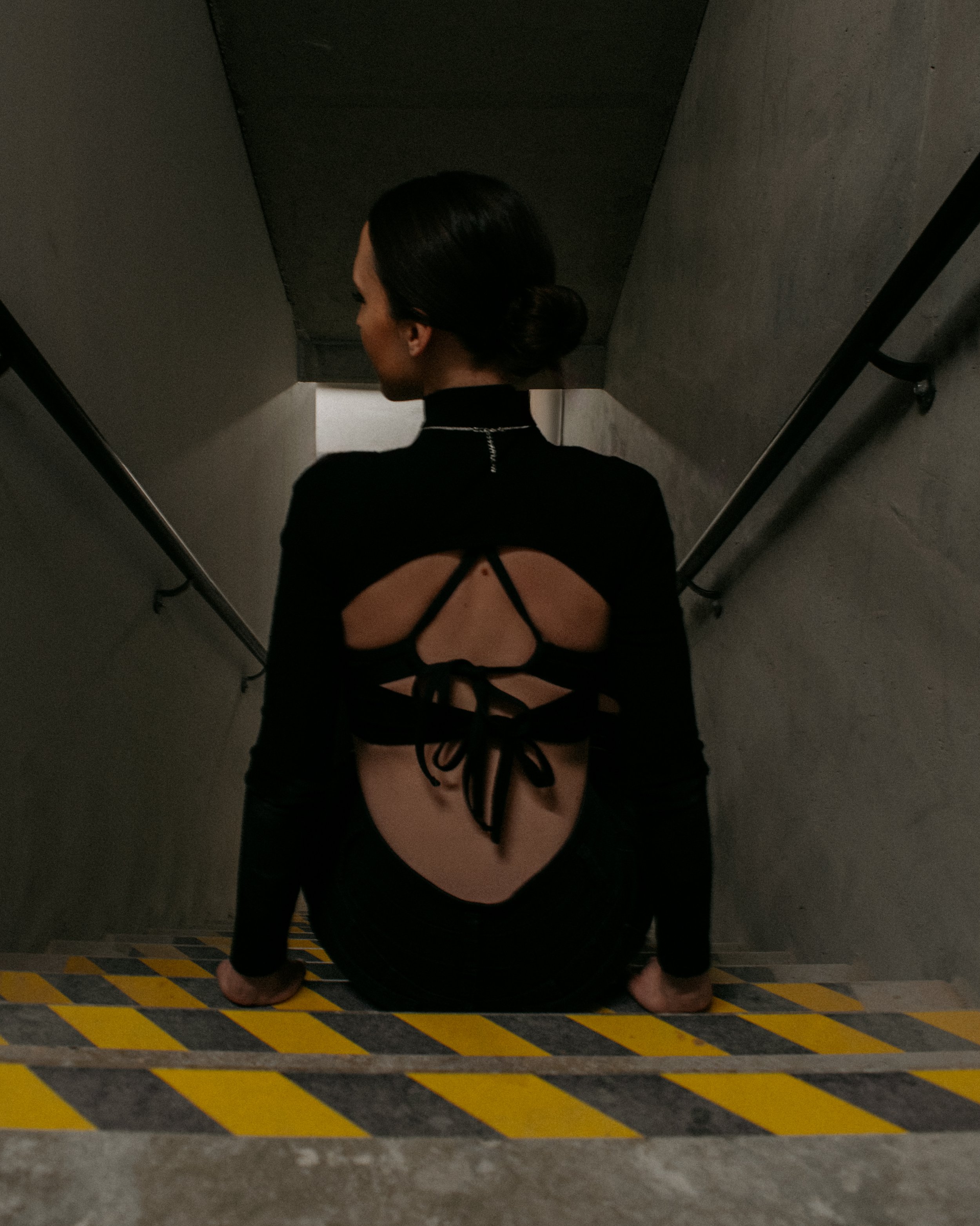 A woman with dark hair styled in a bun, wearing a black dress with an open back and crisscross design, sitting on a staircase with gray walls and yellow and black caution tape on the steps.