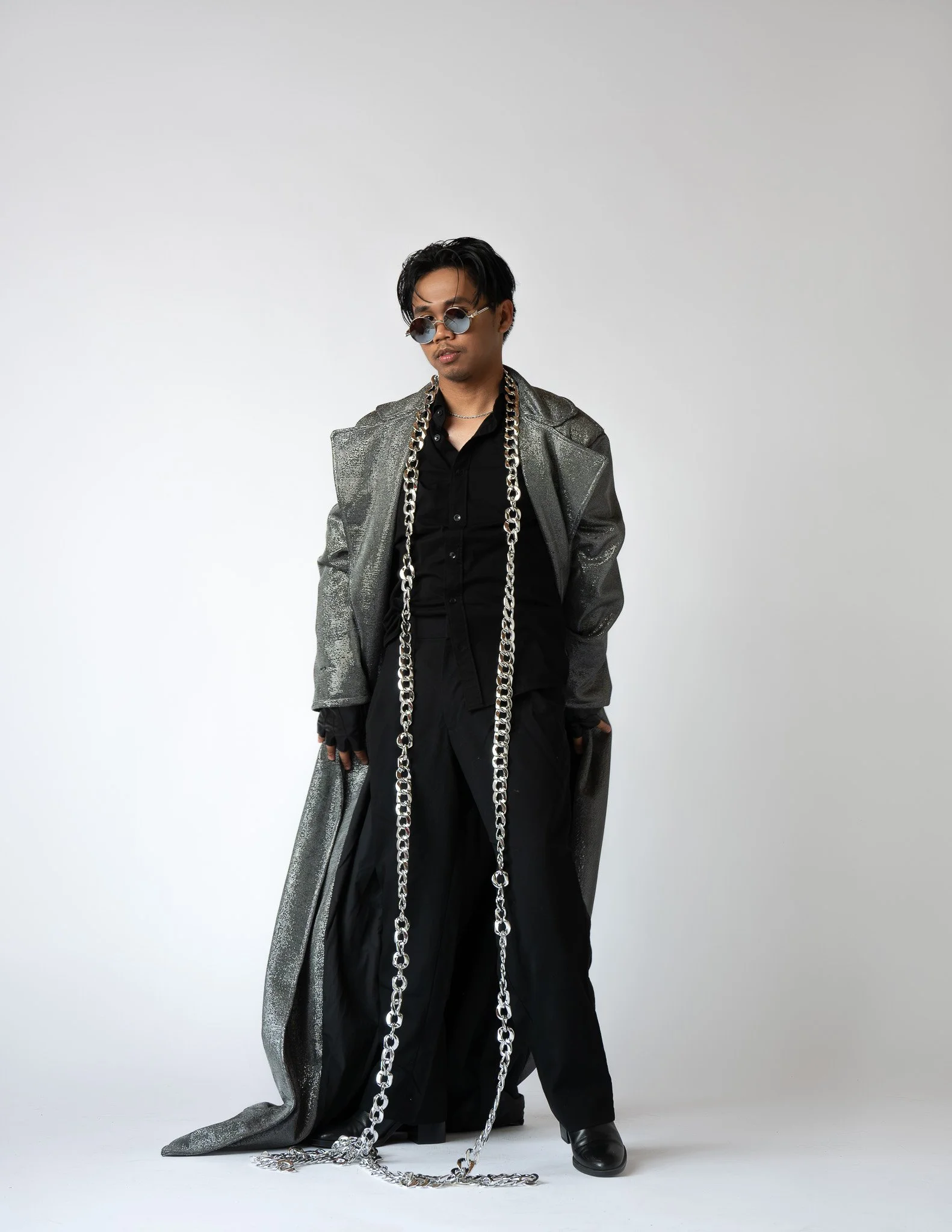 A young man with black hair and sunglasses, wearing a black shirt, silver metallic jacket, and long chains, standing against a plain white background.