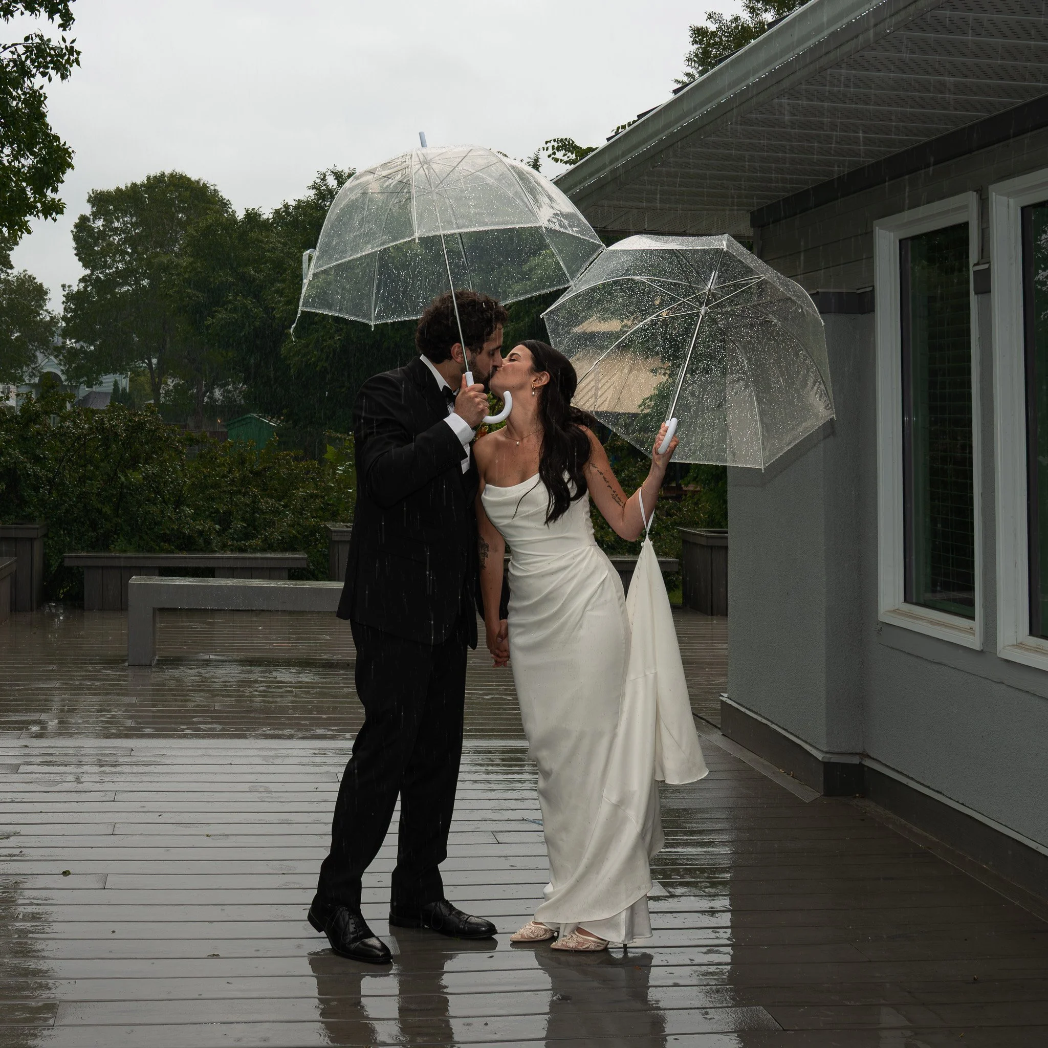 A couple dressed in wedding attire sharing a kiss under umbrellas on a rainy day outside a house.