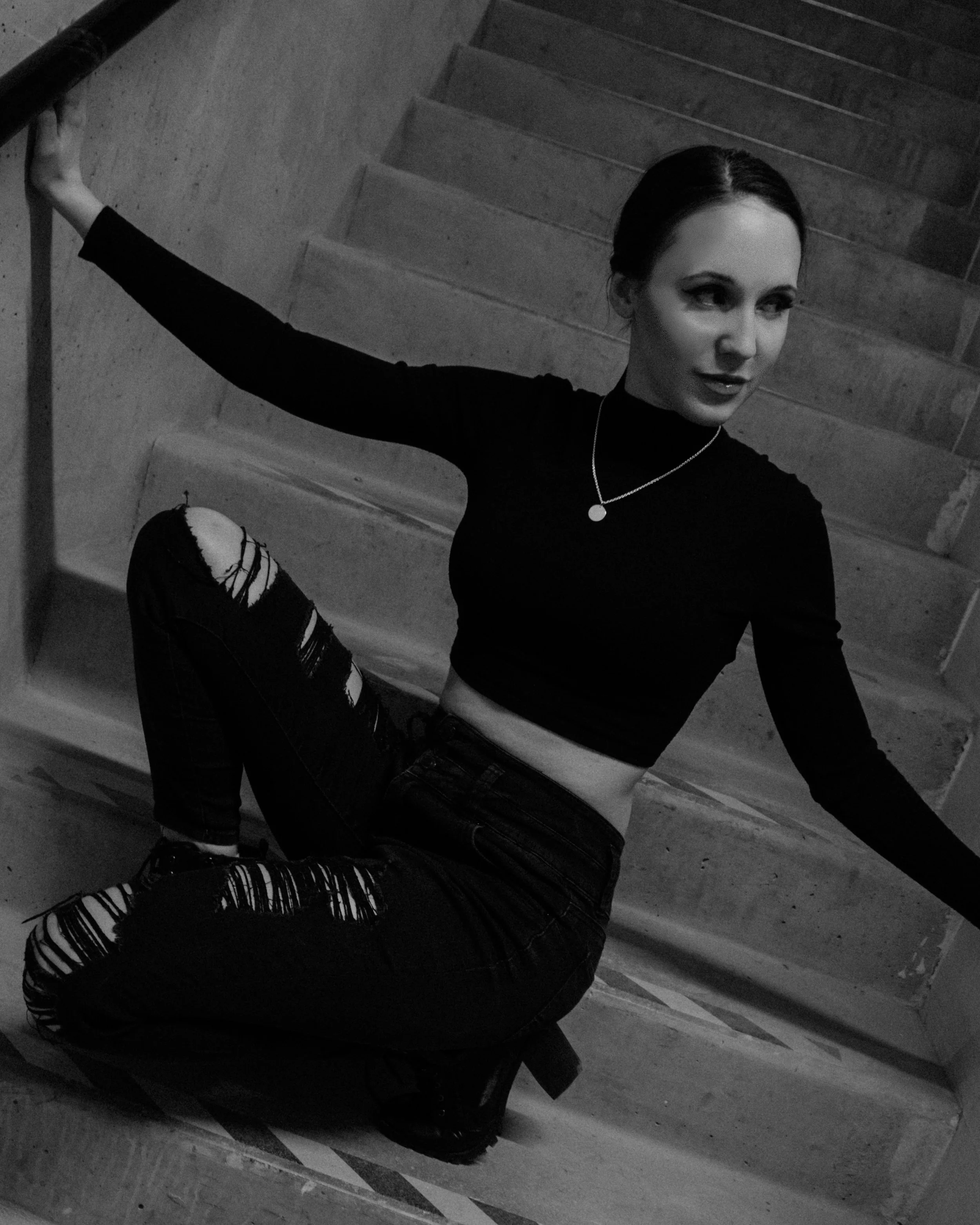 A woman with dark hair in a bun, wearing a black long-sleeve top, ripped jeans, and boots, sitting on a staircase, looking to the side.