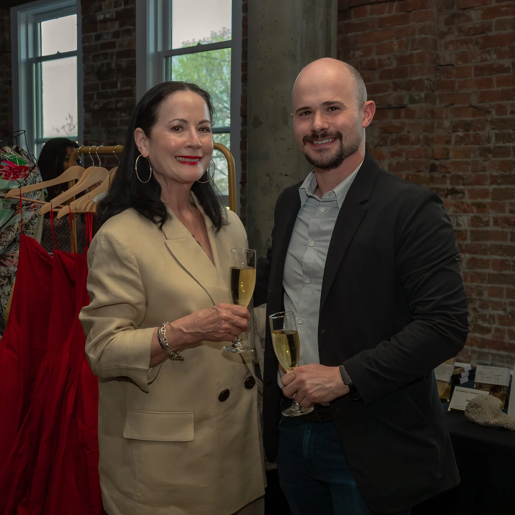 A woman and a man smiling at a social event, holding glasses of champagne, with a brick wall background and clothing racks visible behind them.