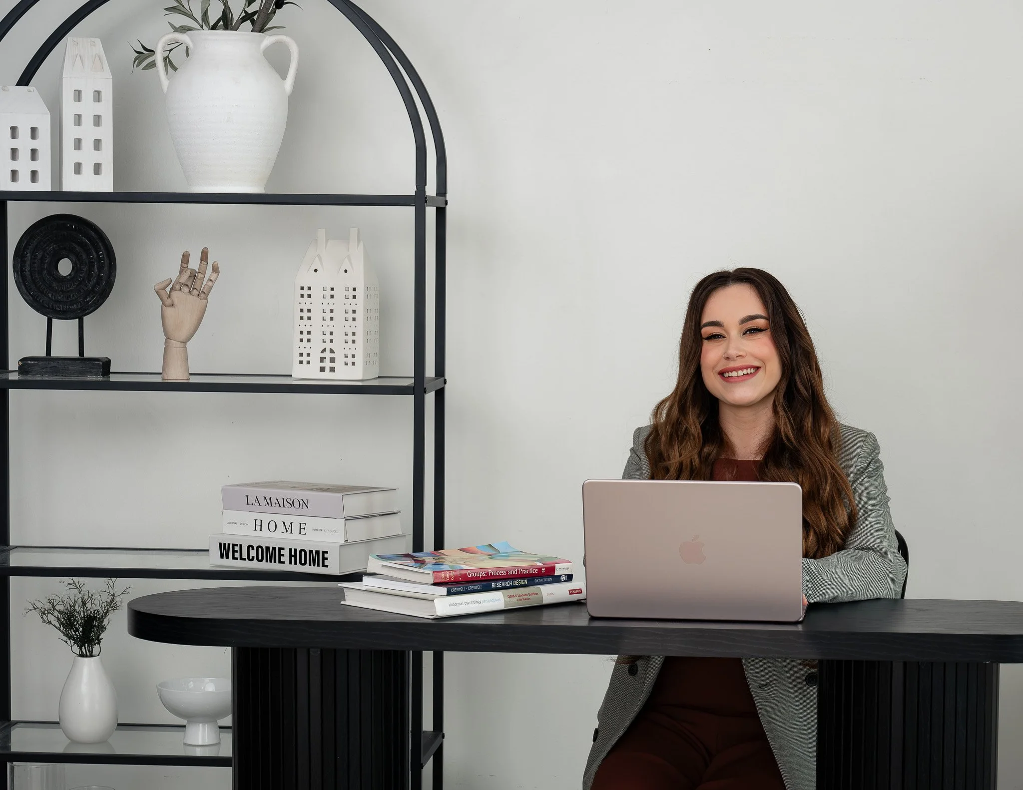 A young woman with long wavy brown hair sitting at a black table with a silver laptop in front of her, in a room with decorative shelves and books behind her.