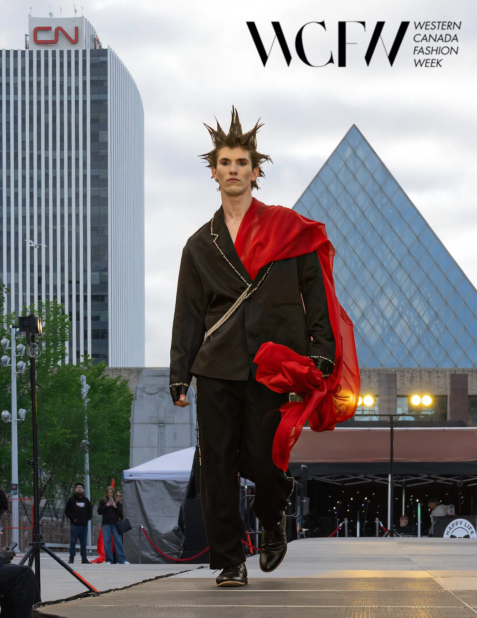 Model walking on stage at Western Canada Fashion Week, cityscape background with modern buildings, overcast sky.