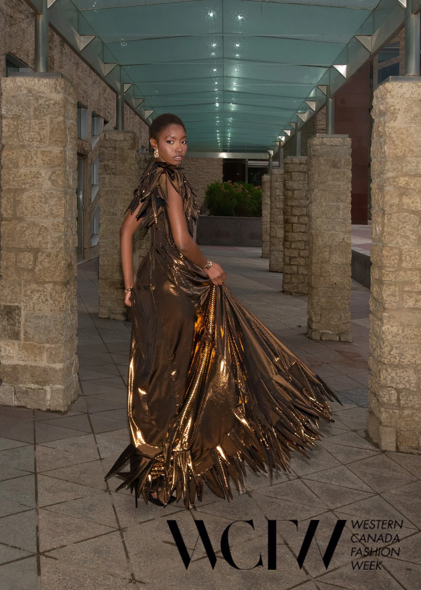 A woman with dark skin and short hair wearing a shiny, metallic gold dress with a ruffled design, standing in a modern outdoor setting with stone columns and a glass ceiling.