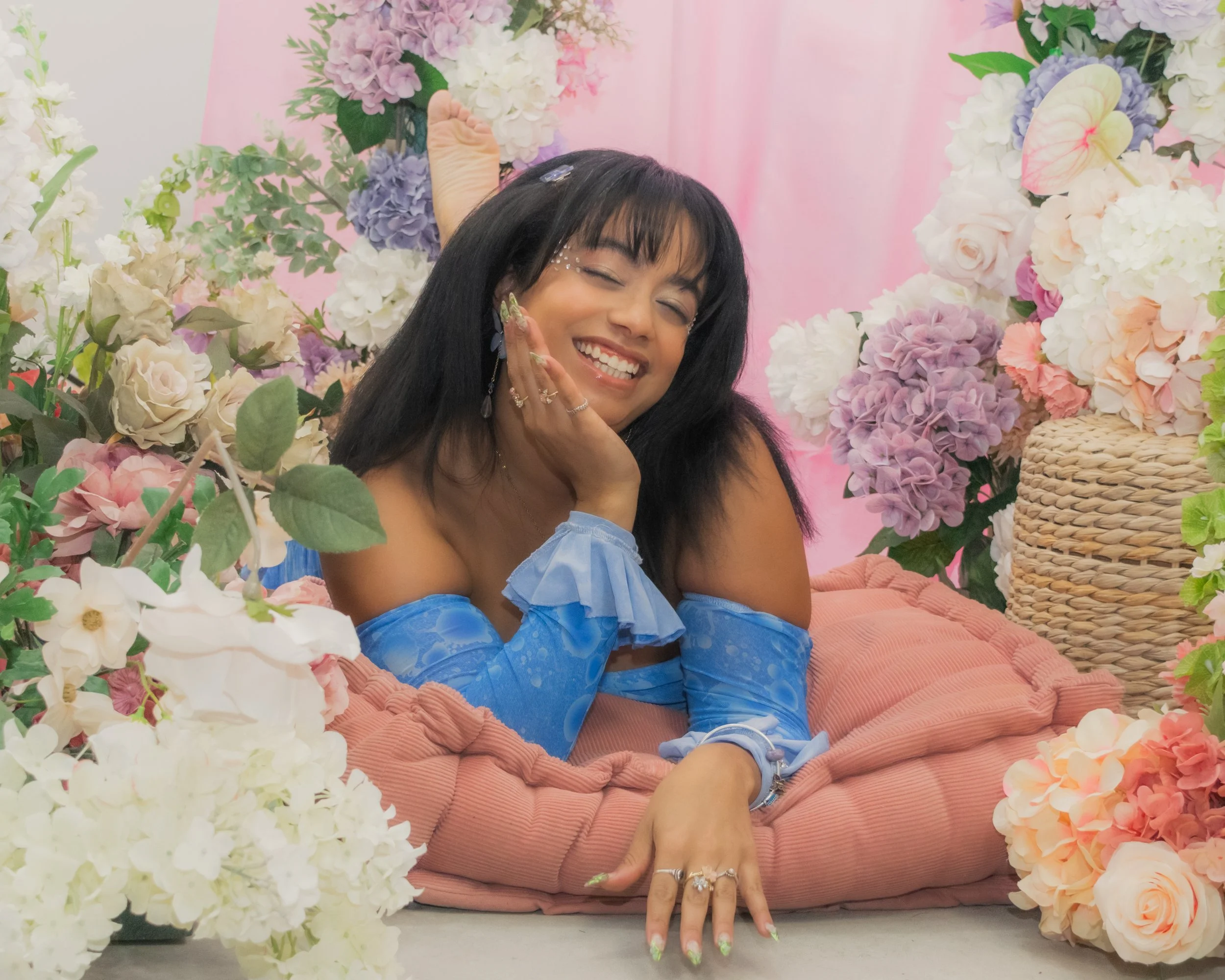 A woman with black hair and a bright smile, wearing blue off-the-shoulder dress with ruffled sleeves, lying on a soft pink cushion surrounded by an abundance of colorful flowers, including hydrangeas and roses, with a pink backdrop.