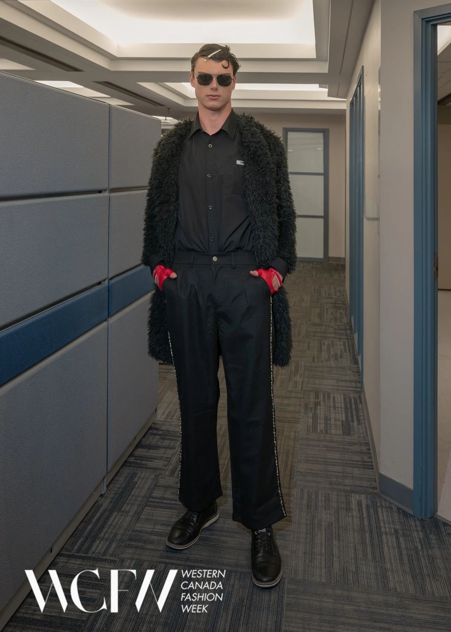 A young male model in black sunglasses, a black shirt, a long black fuzzy coat, red gloves, black loose pants, and black shoes standing in an indoor corridor at Western Canada Fashion Week, with the logo and event name overlayed at the bottom of the 