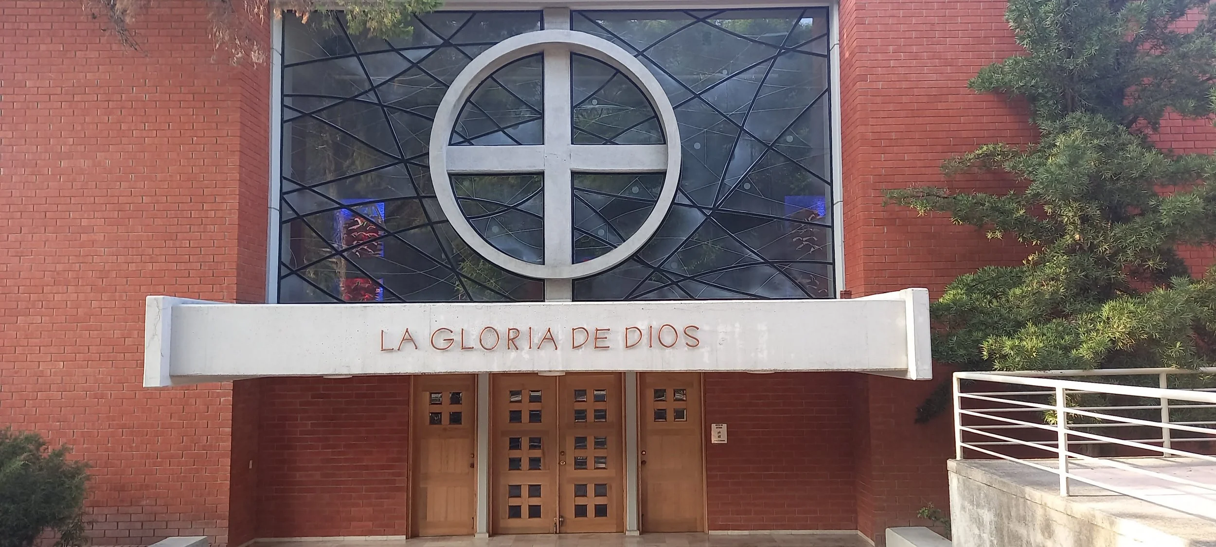 Fachada de una iglesia con puertas de madera, un letrero que dice 'La Gloria de Dios', una cruz estilizada en el ventanal de cristal y árboles a los lados.