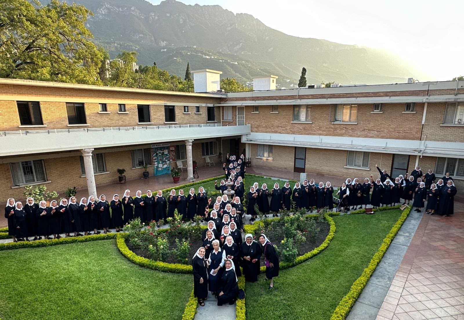 Grupo de monjas con hábitos en un jardín de un edificio, con montañas al fondo, durante el atardecer.