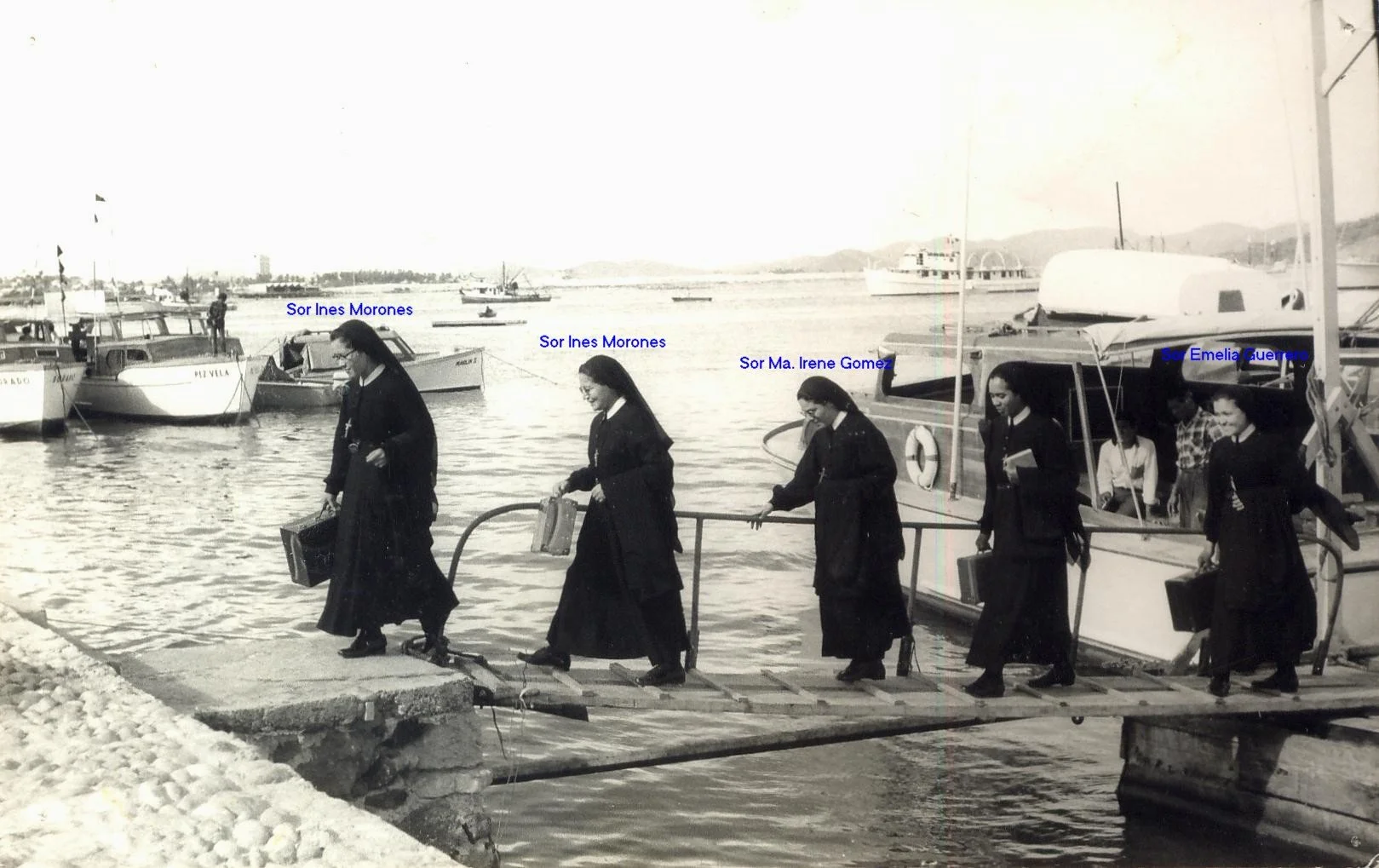 Cinco monjas bajando de un muelle hacia el agua, con barcos en el puerto de fondo, en una fotografía en blanco y negro.